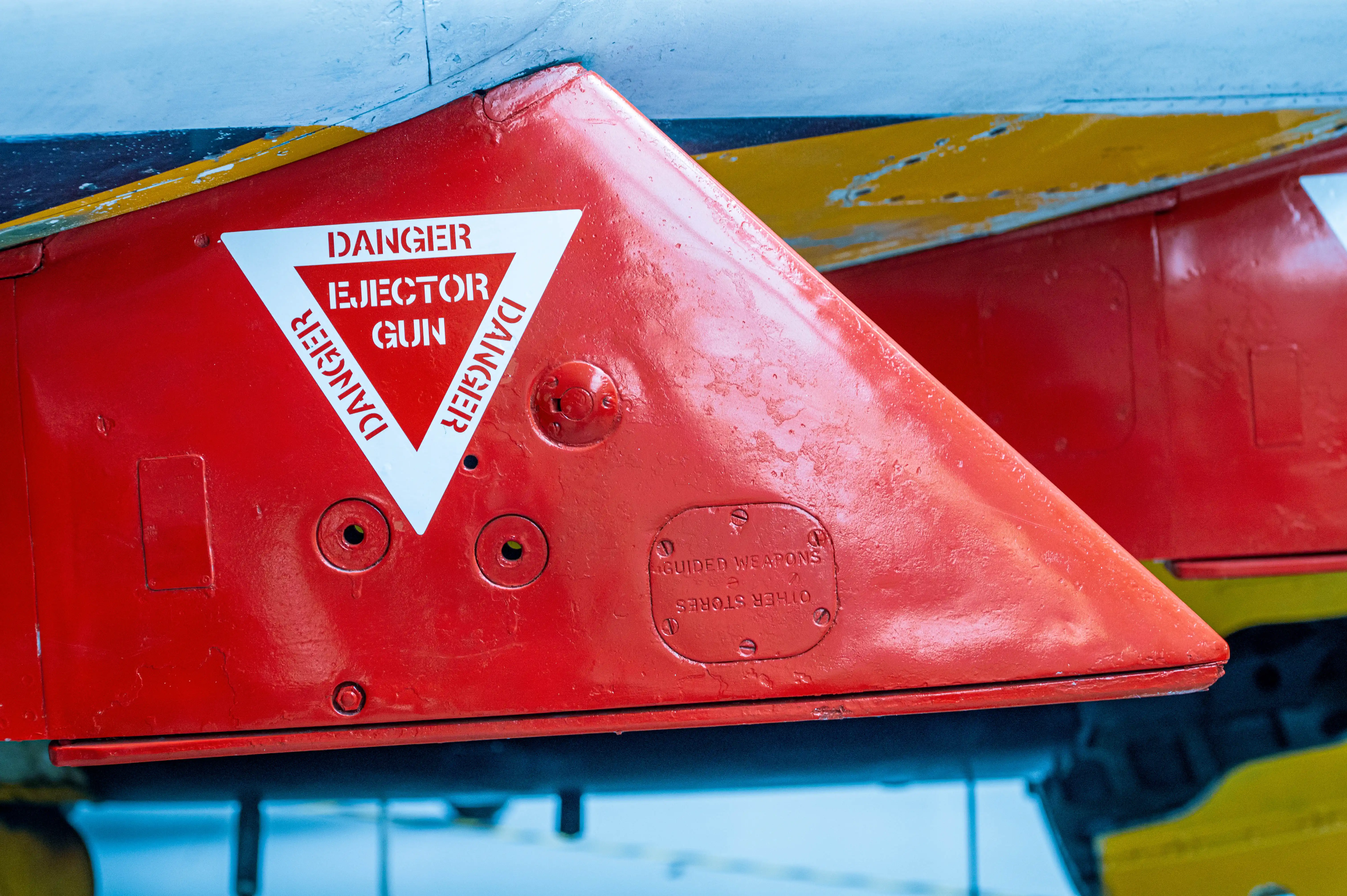 A red triangular warning label on an aircraft states 'DANGER EJECTOR GUN' and 'DANGER' written vertically on the sides. Below the triangle, a plate reads 'OTHER STORES GUIDED WEAPONS'.