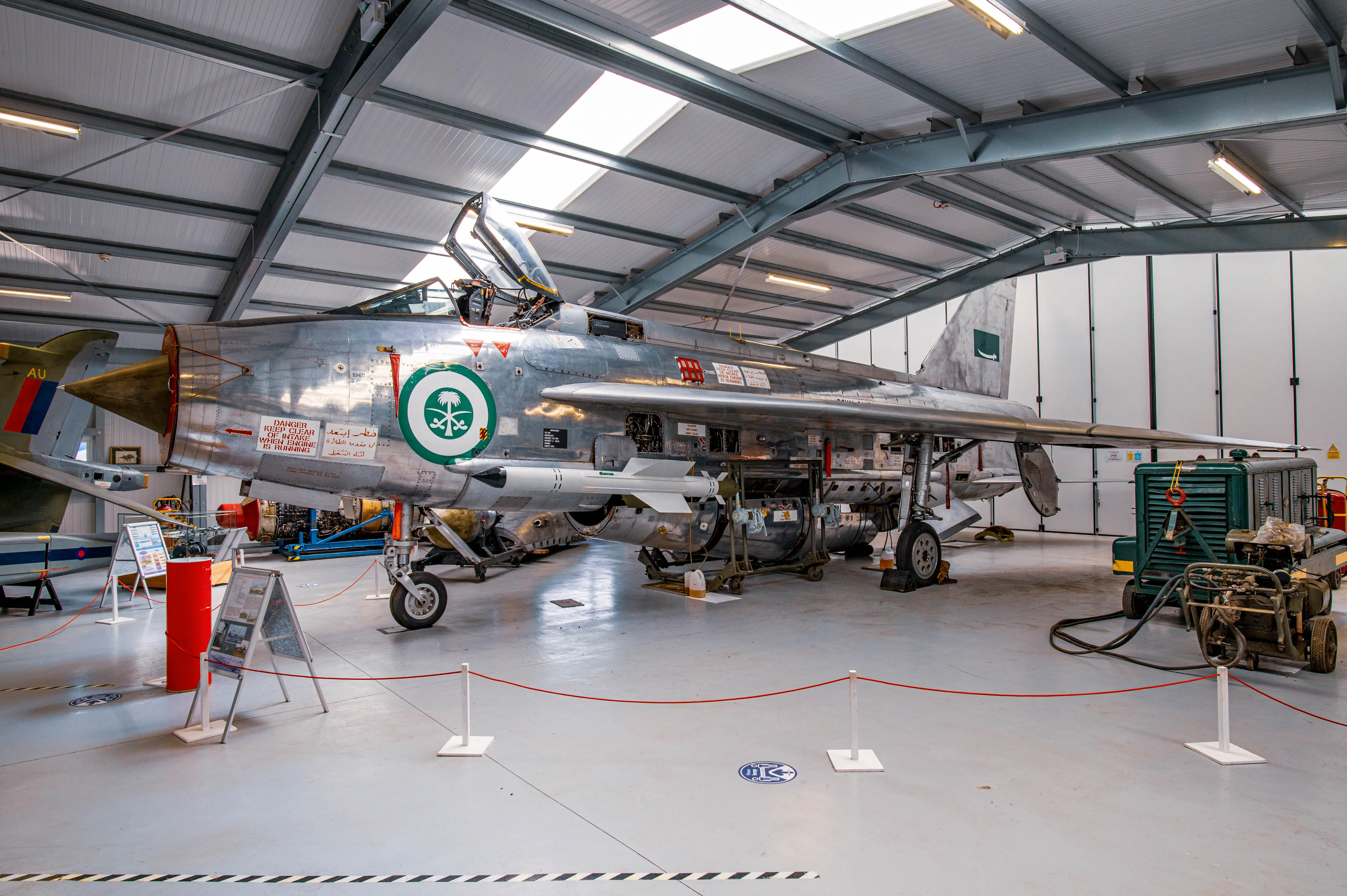 A silver English Electric Lightning fighter jet, emblazoned with the Saudi Arabian emblem, stands on display in a museum hangar. The cockpit canopy is open, revealing intricate details of the aircraft's interior. A missile is attached beneath its wing. In the background, other aircraft and museum elements, including informational stands and a ground power unit, can be seen.