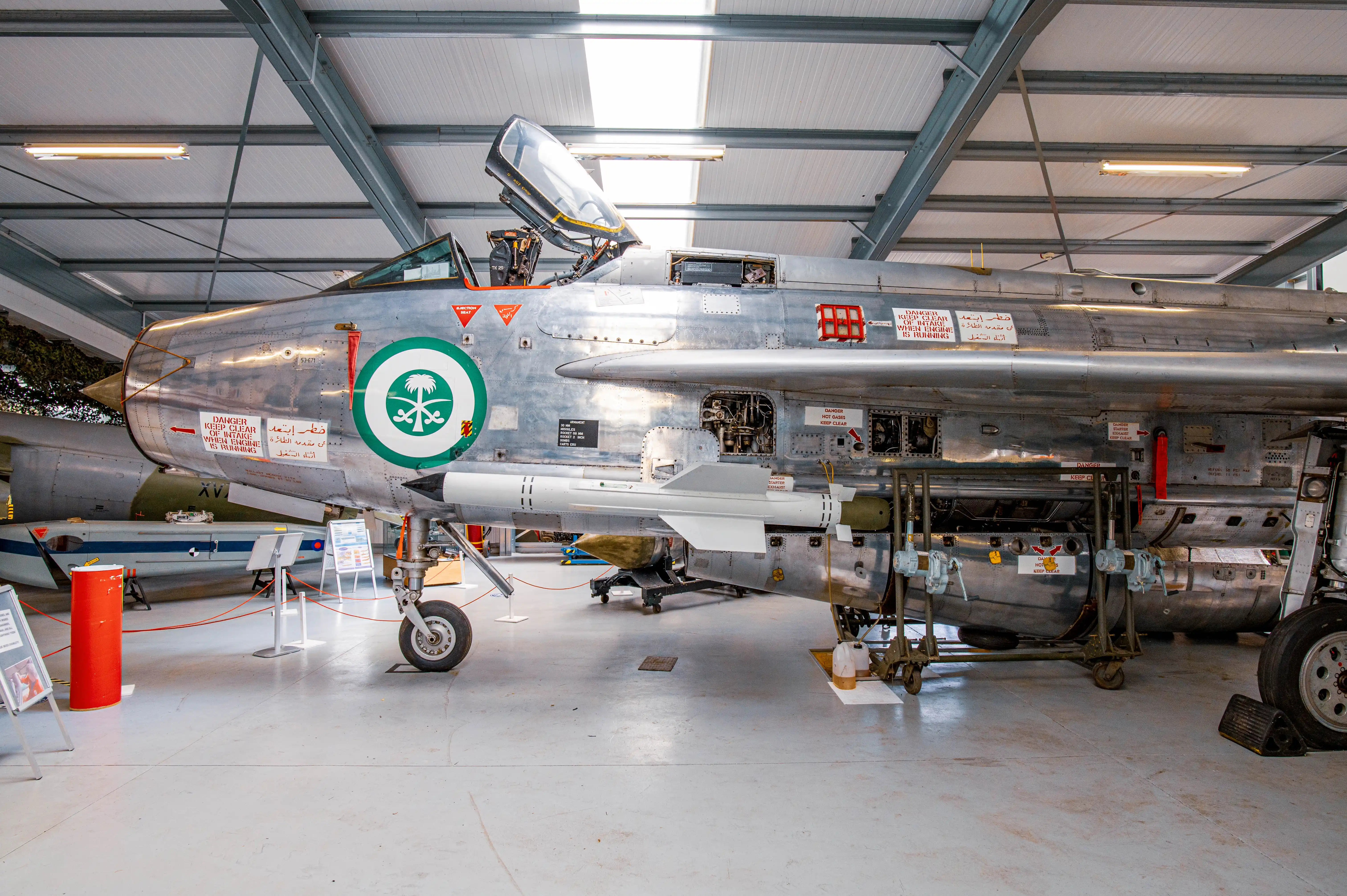 A side profile of a silver fighter jet with the Saudi Arabian emblem displayed in a museum. The cockpit is open, and a missile is attached beneath the wing.