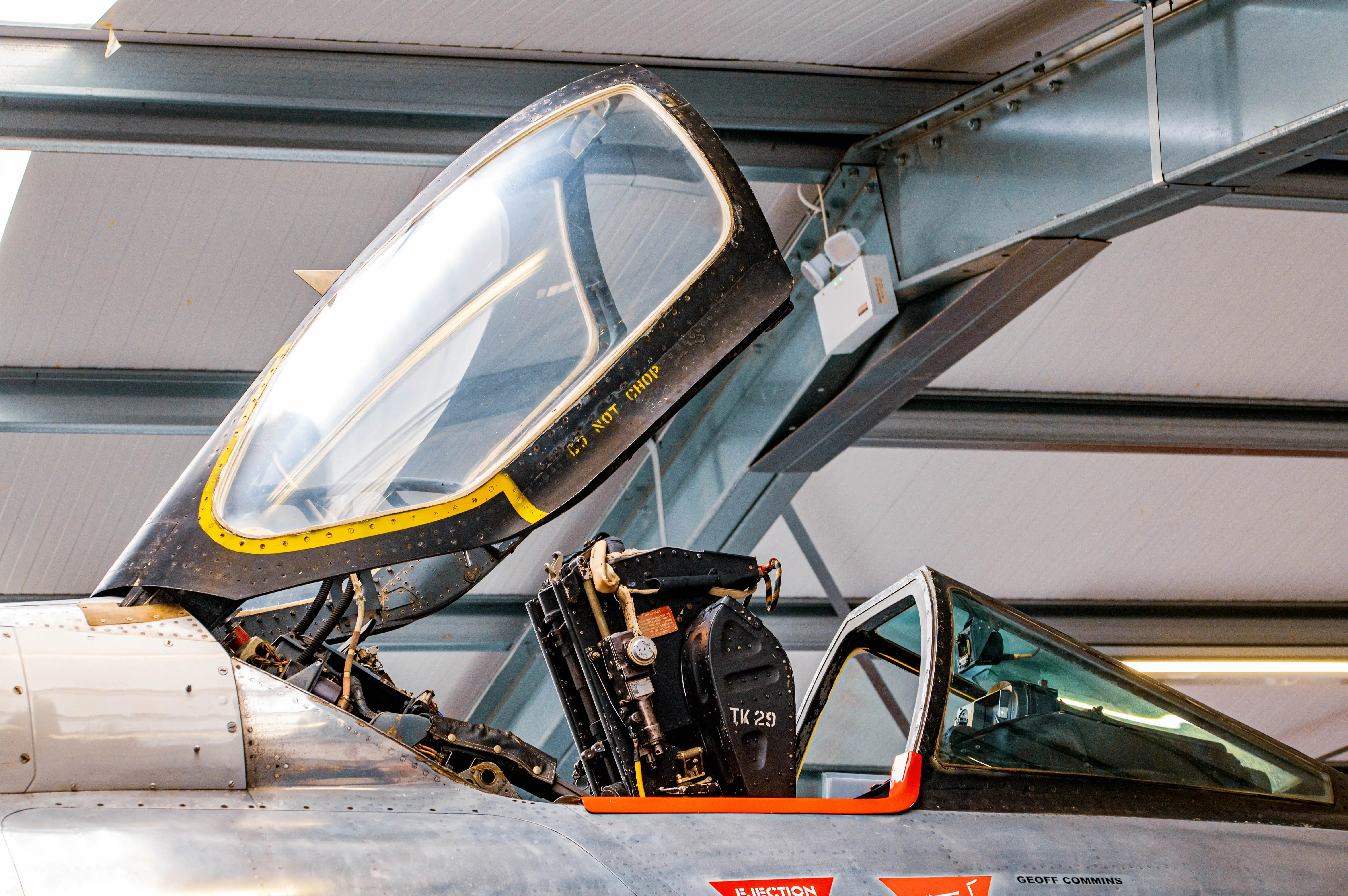 A close-up photograph showing the open cockpit of a vintage fighter jet. The pilot's seat with its ejection system is prominent, along with the instrument panel and canopy frame. Warning labels are visible on the canopy and near the ejection seat.