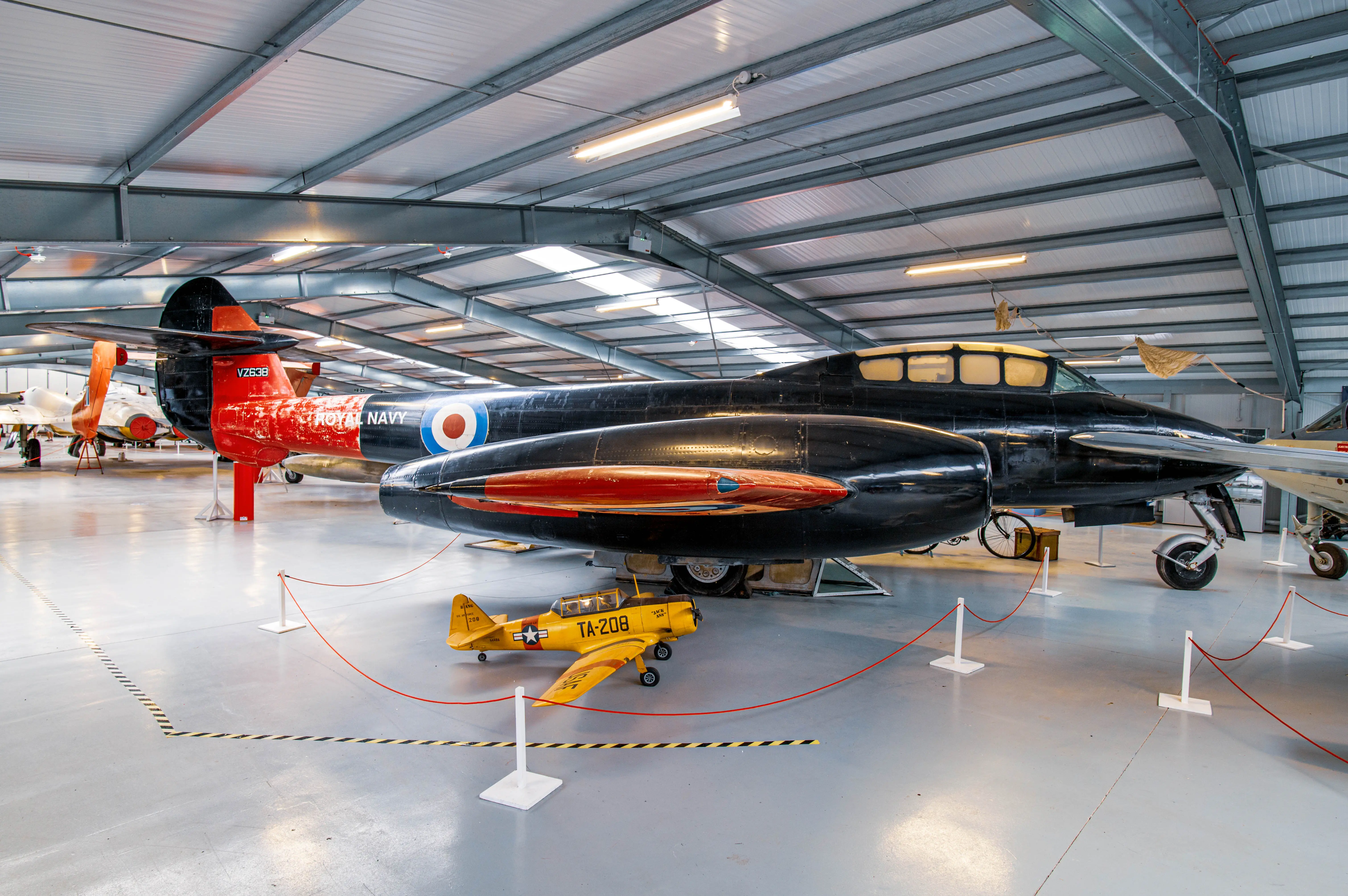 A large, black Royal Navy jet aircraft and a small yellow model aircraft are parked inside a brightly lit aircraft hangar