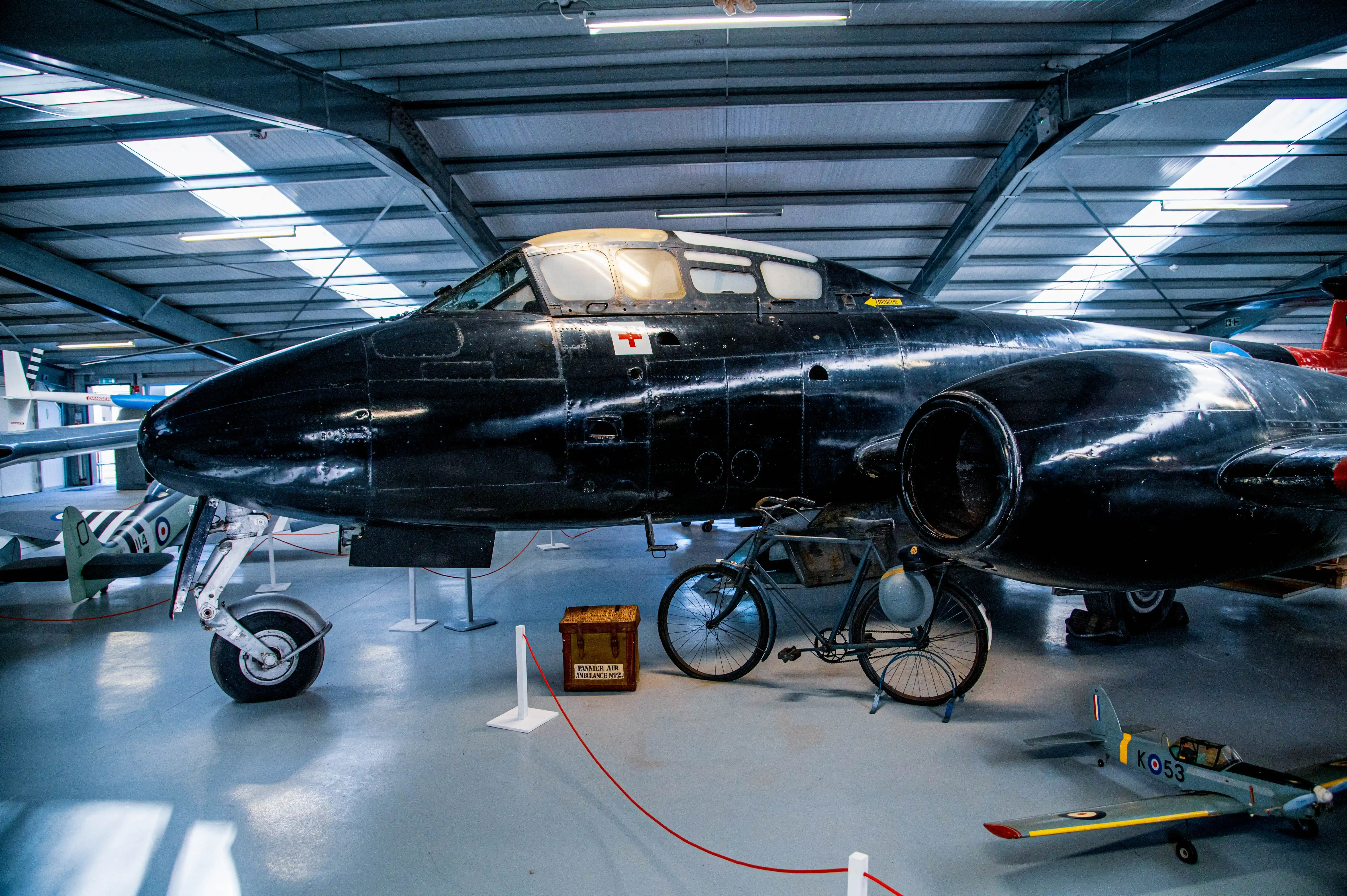 A Gloster Meteor jet aircraft with a black glossy finish is parked inside a hangar. A vintage grey bicycle stands next to it, and a small model plane is visible in the foreground.