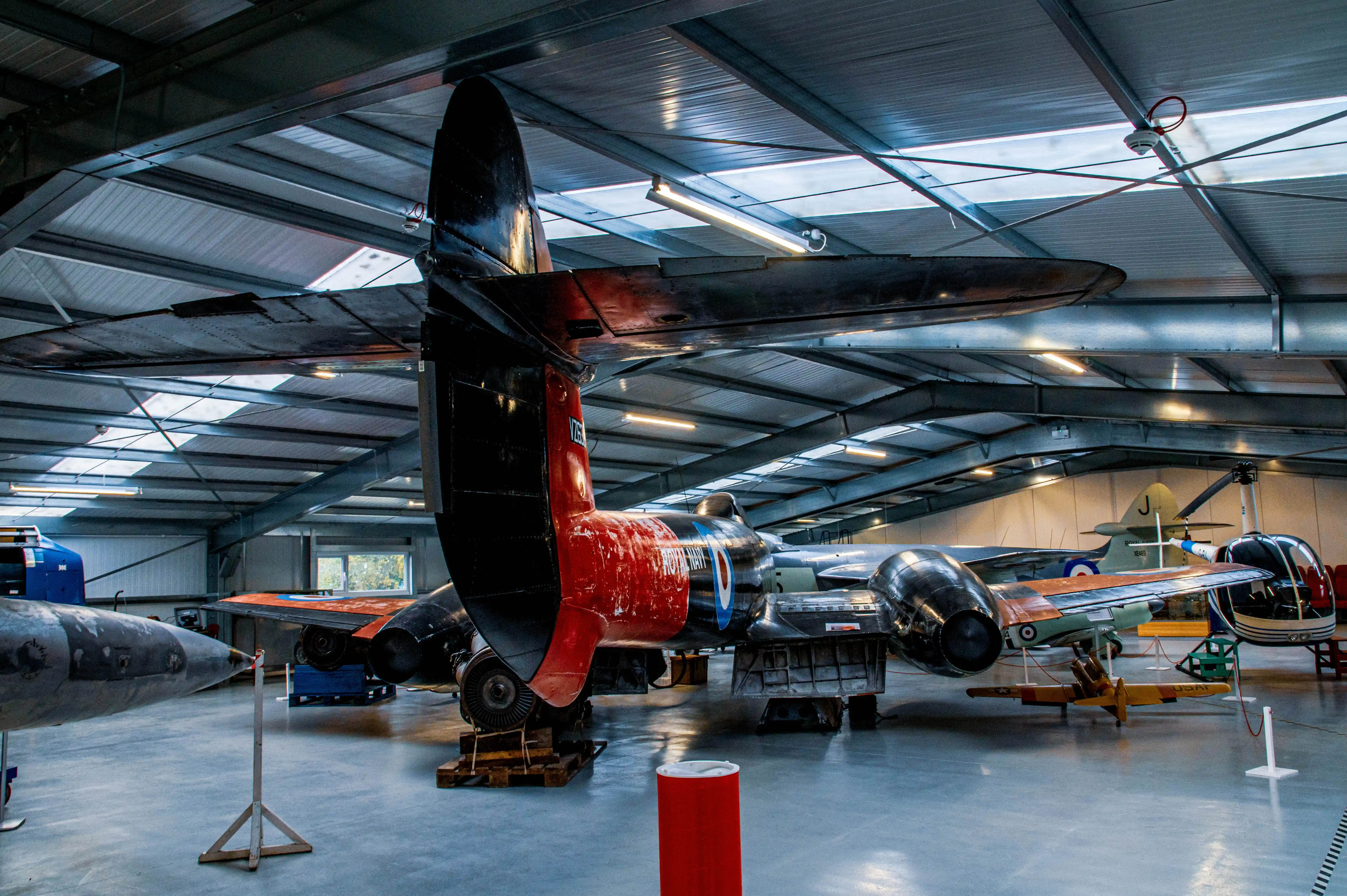 Rear view of a vintage Royal Navy jet trainer aircraft with red and black tail fin, parked inside a museum hangar. Other aircraft and display items are visible in the background under a corrugated metal roof with skylights.