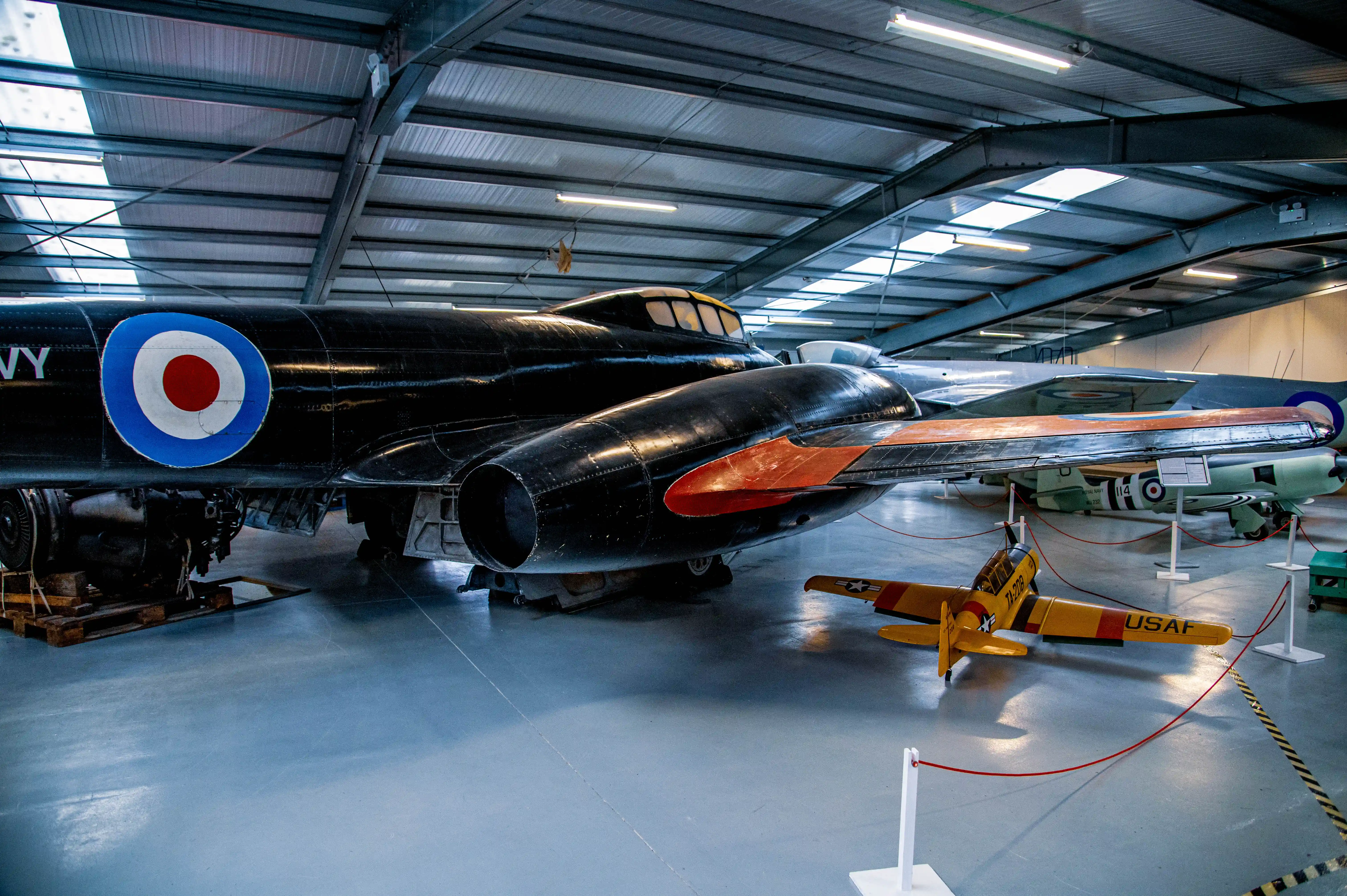 Interior view of a museum hangar displaying various vintage aircraft. In the foreground is a dark-coloured jet with a British Navy roundel, and a smaller yellow training aircraft with 