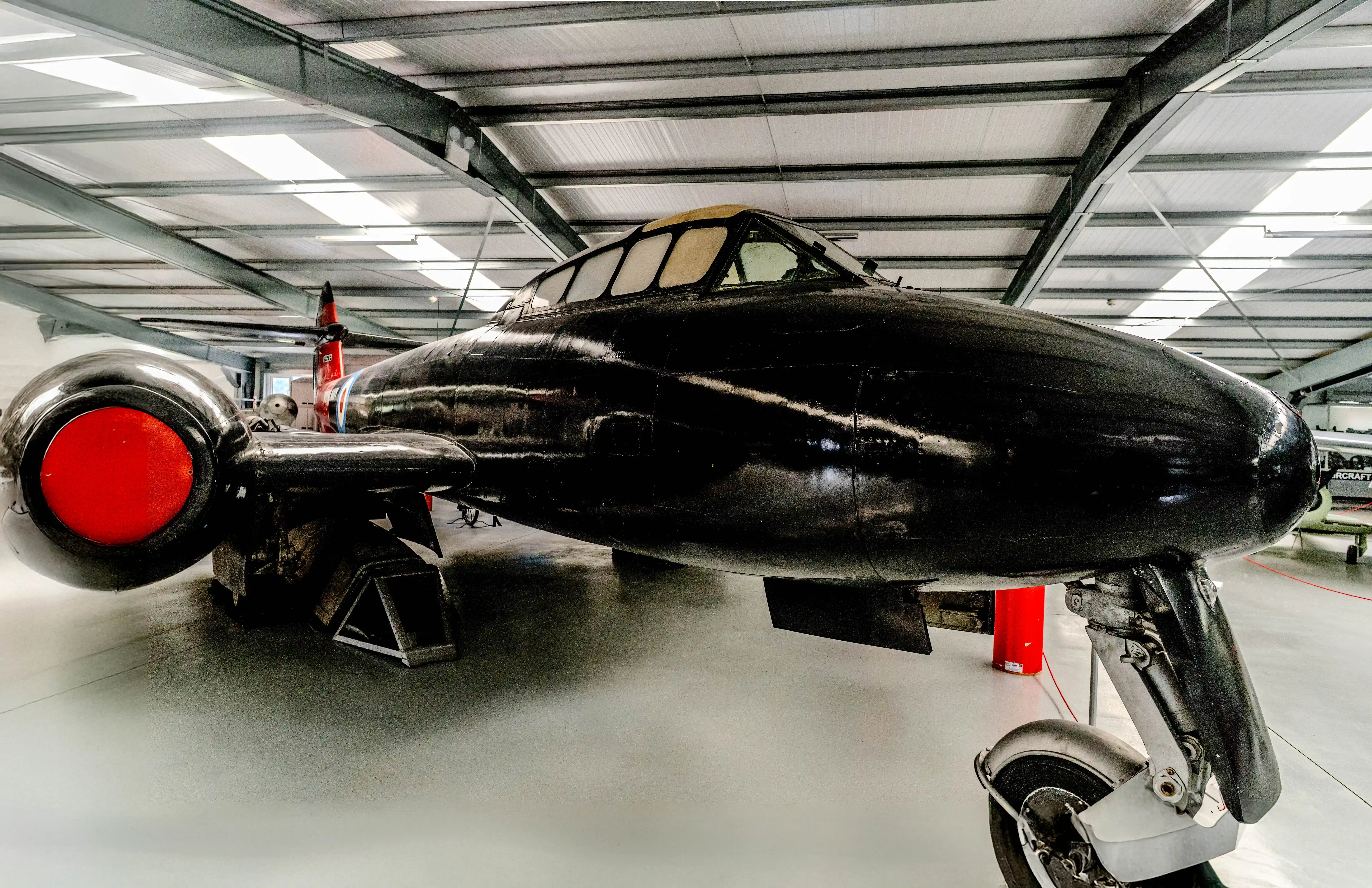 A close-up, side-profile view of a glossy black jet aircraft with prominent red engine intakes, parked inside a well-lit hangar with a corrugated roof.