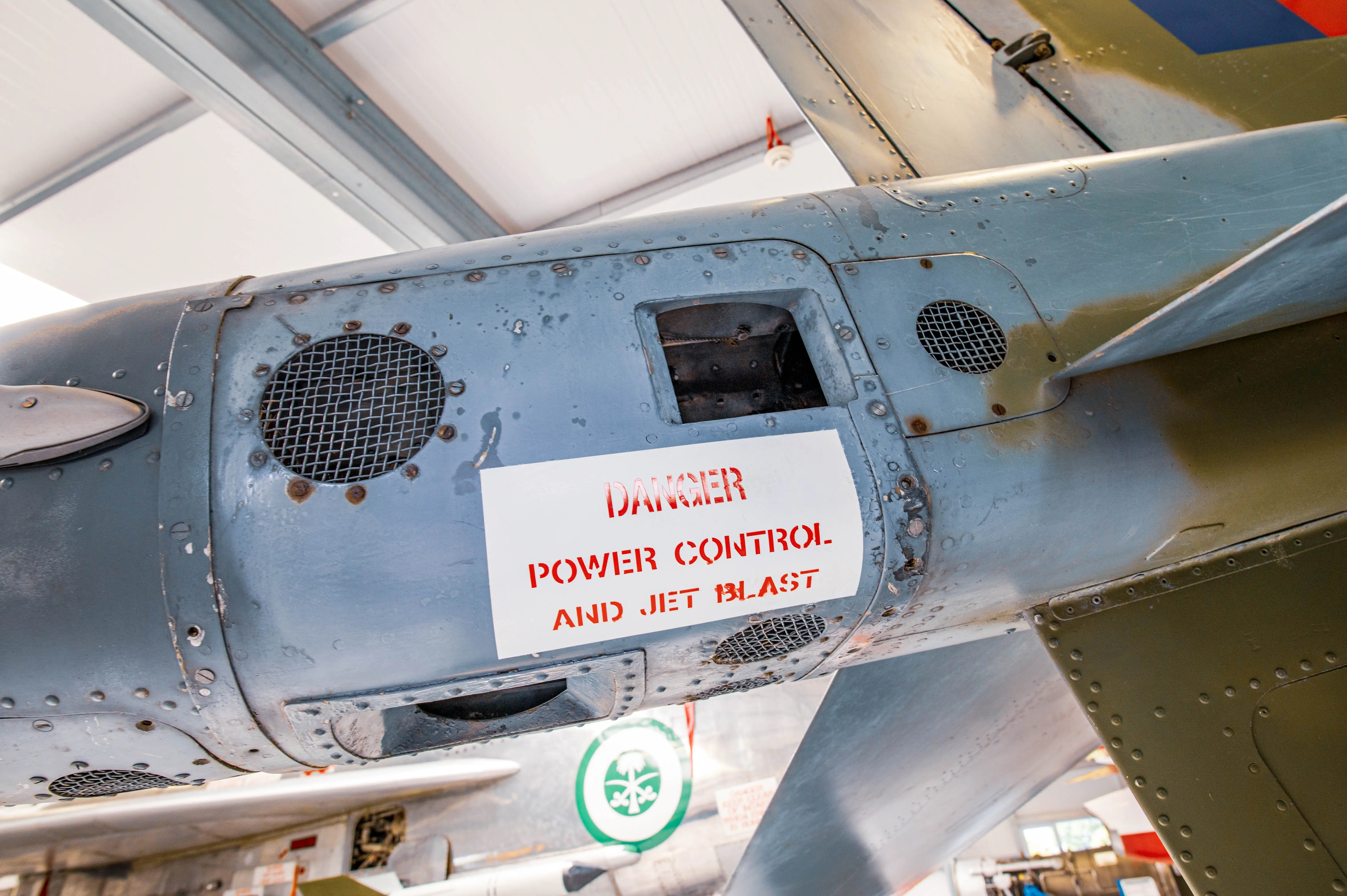 A white rectangular sign with red lettering reading DANGER POWER CONTROL AND JET BLAST is affixed to the grey fuselage of an aircraft.