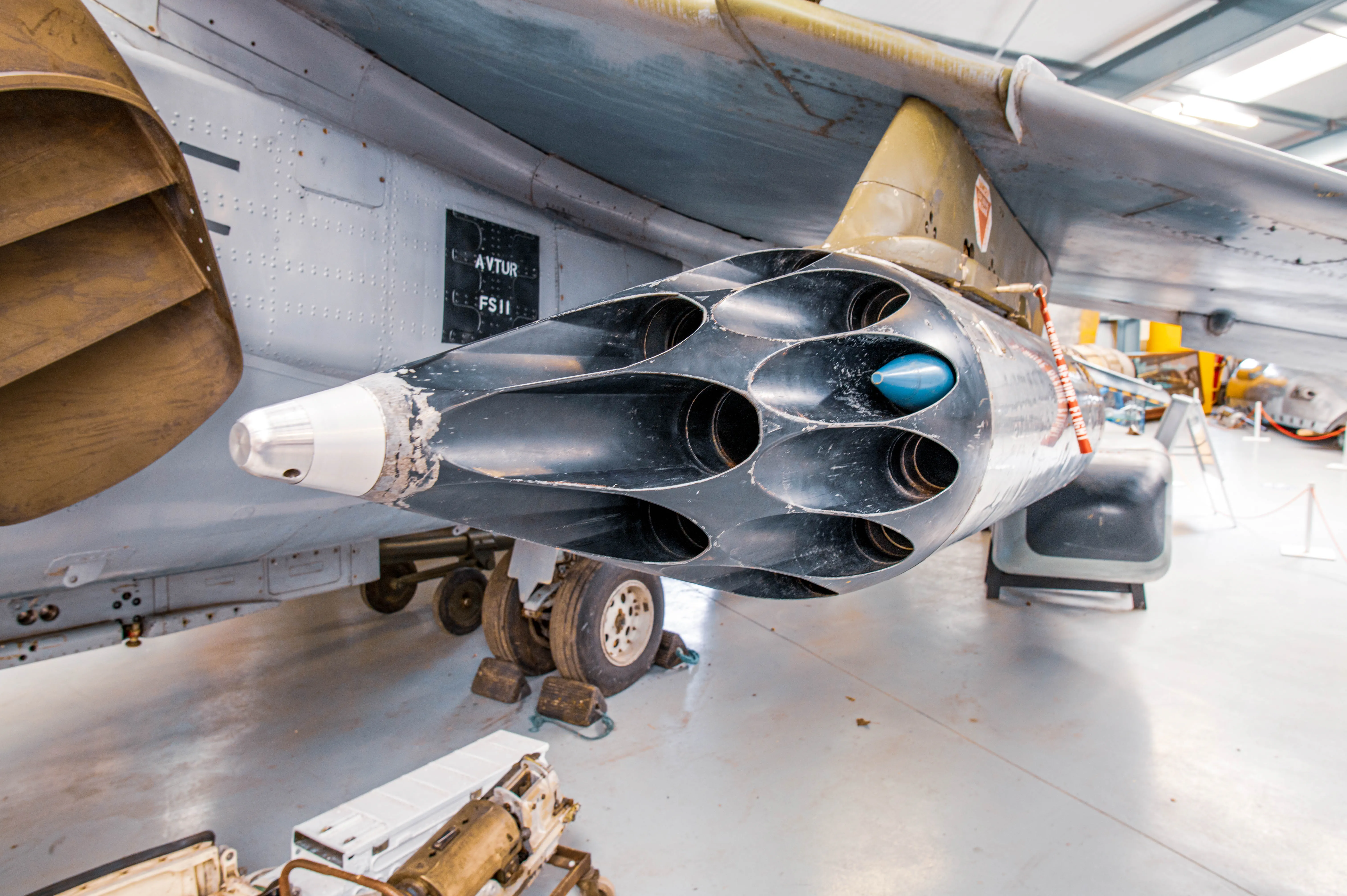 A close-up shot of a black, multi-barrelled rocket launcher mounted under a grey aircraft wing. A single blue-tipped rocket is loaded into the launcher. The aircraft's landing gear and part of its engine are also in view.