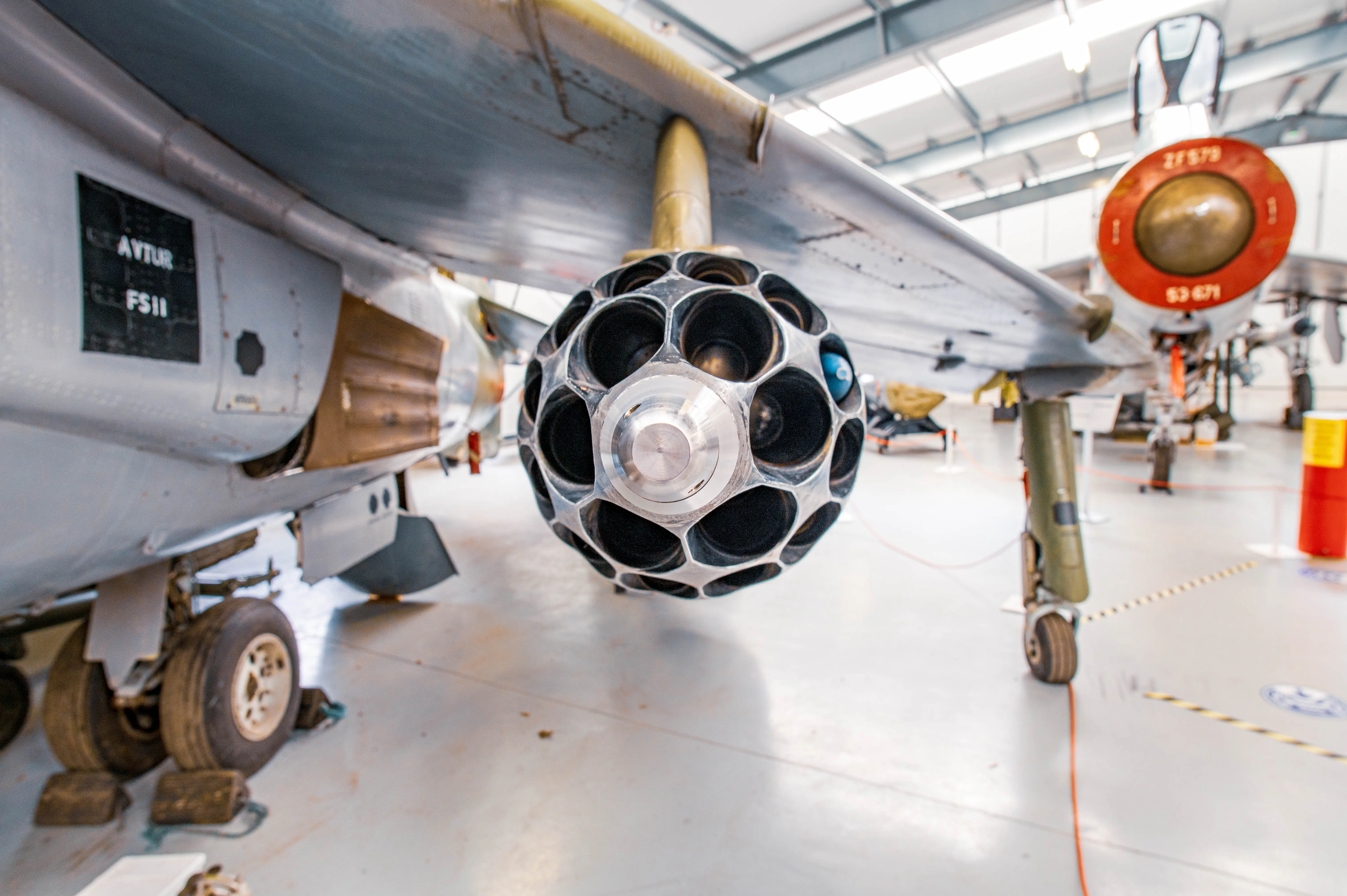 Close-up side view of a hexagonal rocket pod attached to the underside of a grey aircraft wing. The pod is metallic with a series of dark openings and a silver nose cone. The aircraft's landing gear and a blurred background of a hangar are visible.