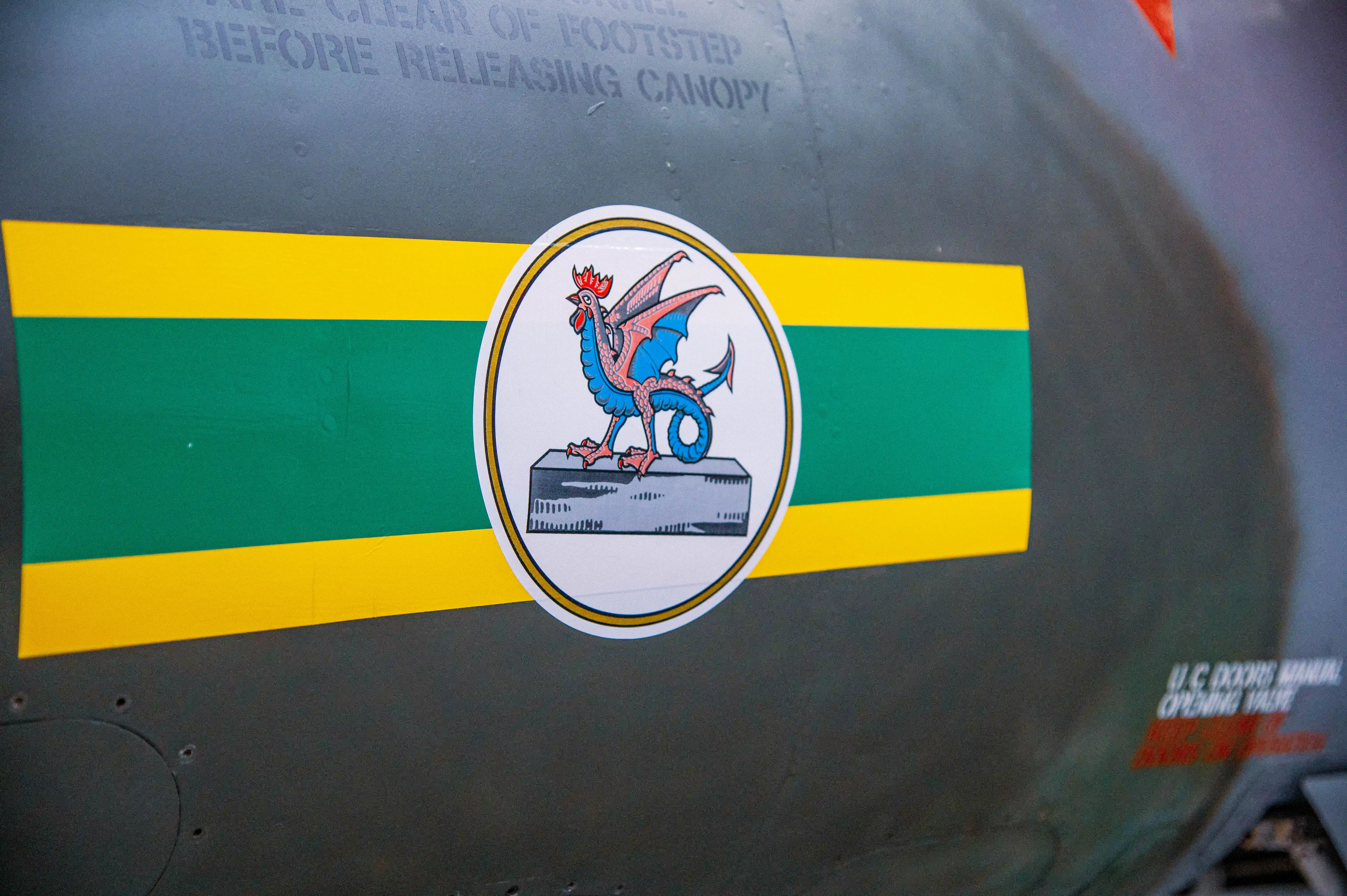 Close-up of a grey aircraft rocket pod with multiple oval-shaped openings, mounted beneath a wing. A red REMOVE BEFORE FLIGHT tag is visible.