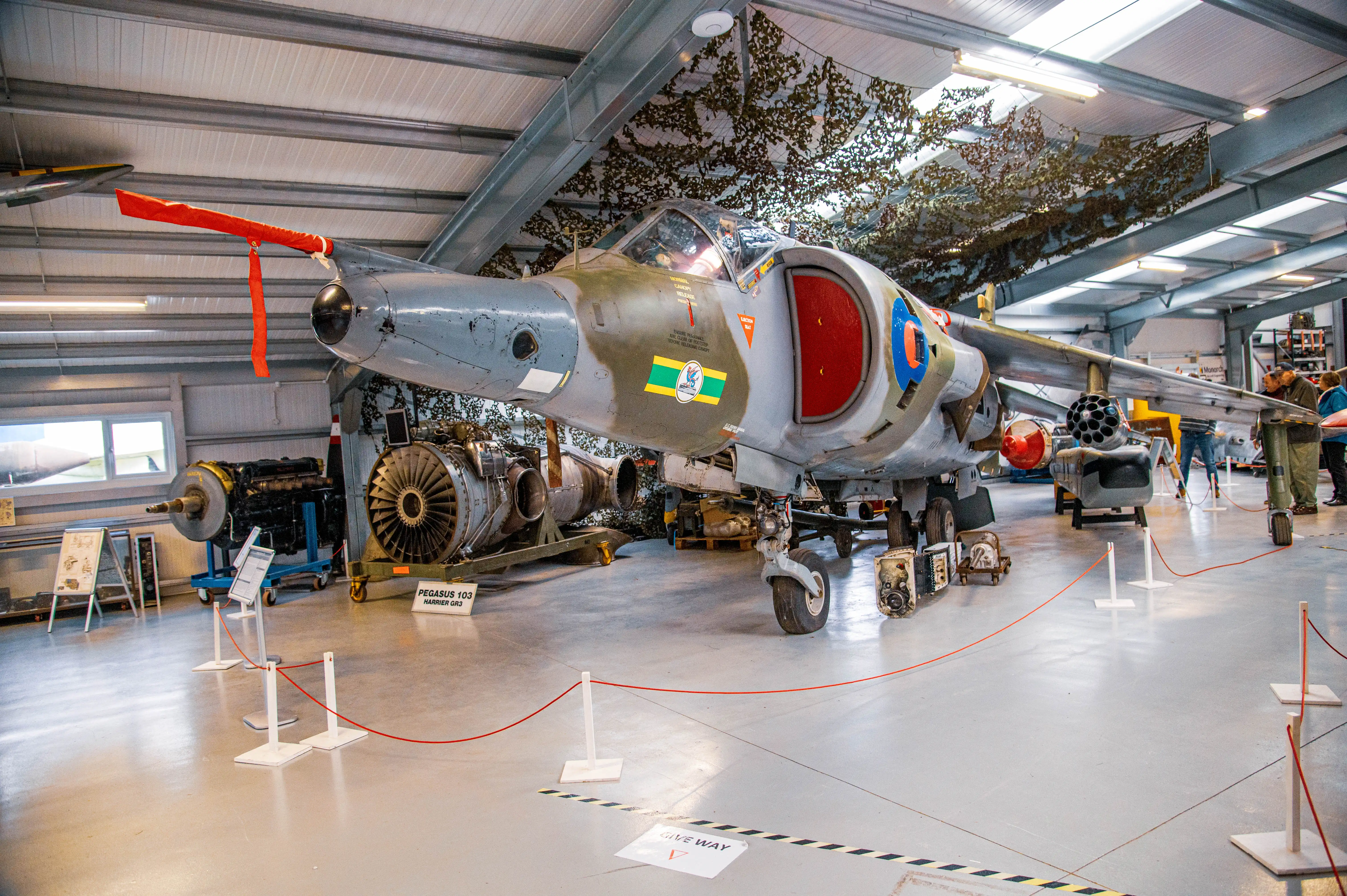 A Harrier GR3 jet aircraft, painted in camouflage colours with red and blue markings, sits in a museum hangar. Display ropes guide visitors around the jet, which is positioned next to a Pegasus engine.