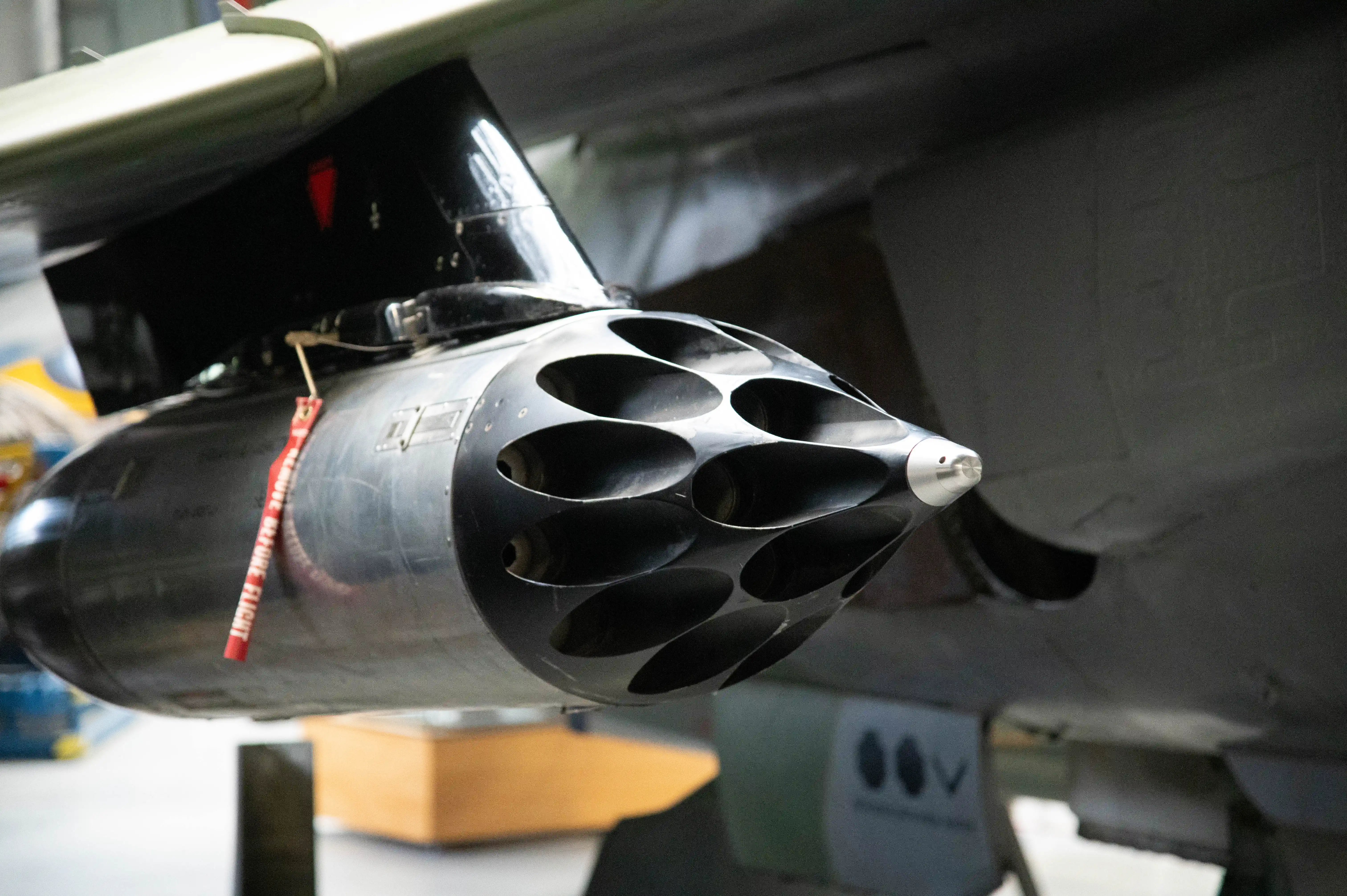 Close-up of a grey aircraft rocket pod with multiple oval-shaped openings, mounted beneath a wing. A red 