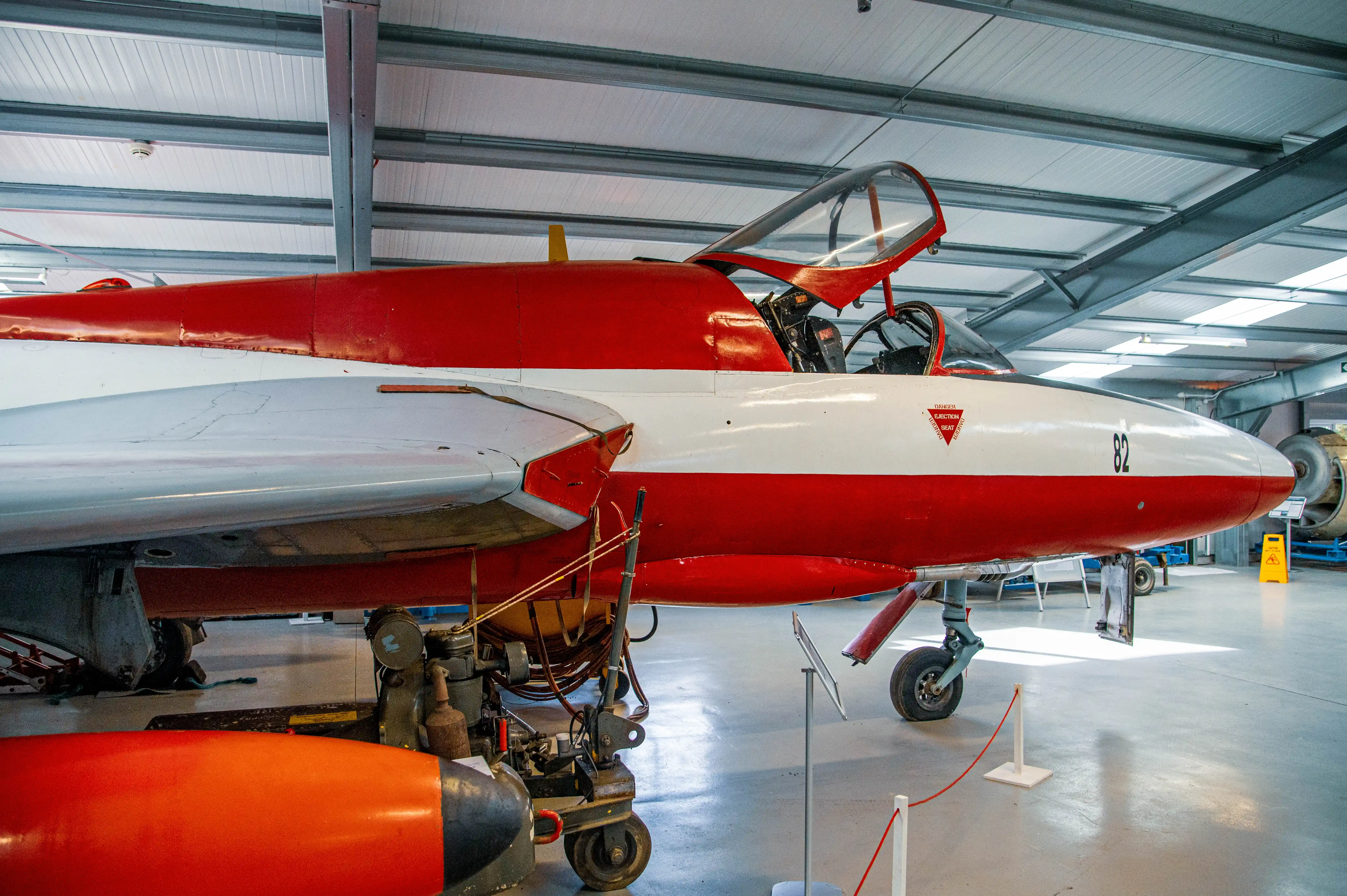 Close-up side view of a vintage red and white jet aircraft parked inside a museum or hangar. The cockpit is open, revealing the pilot's seat. A warning sticker about the ejection seat is visible on the fuselage.
