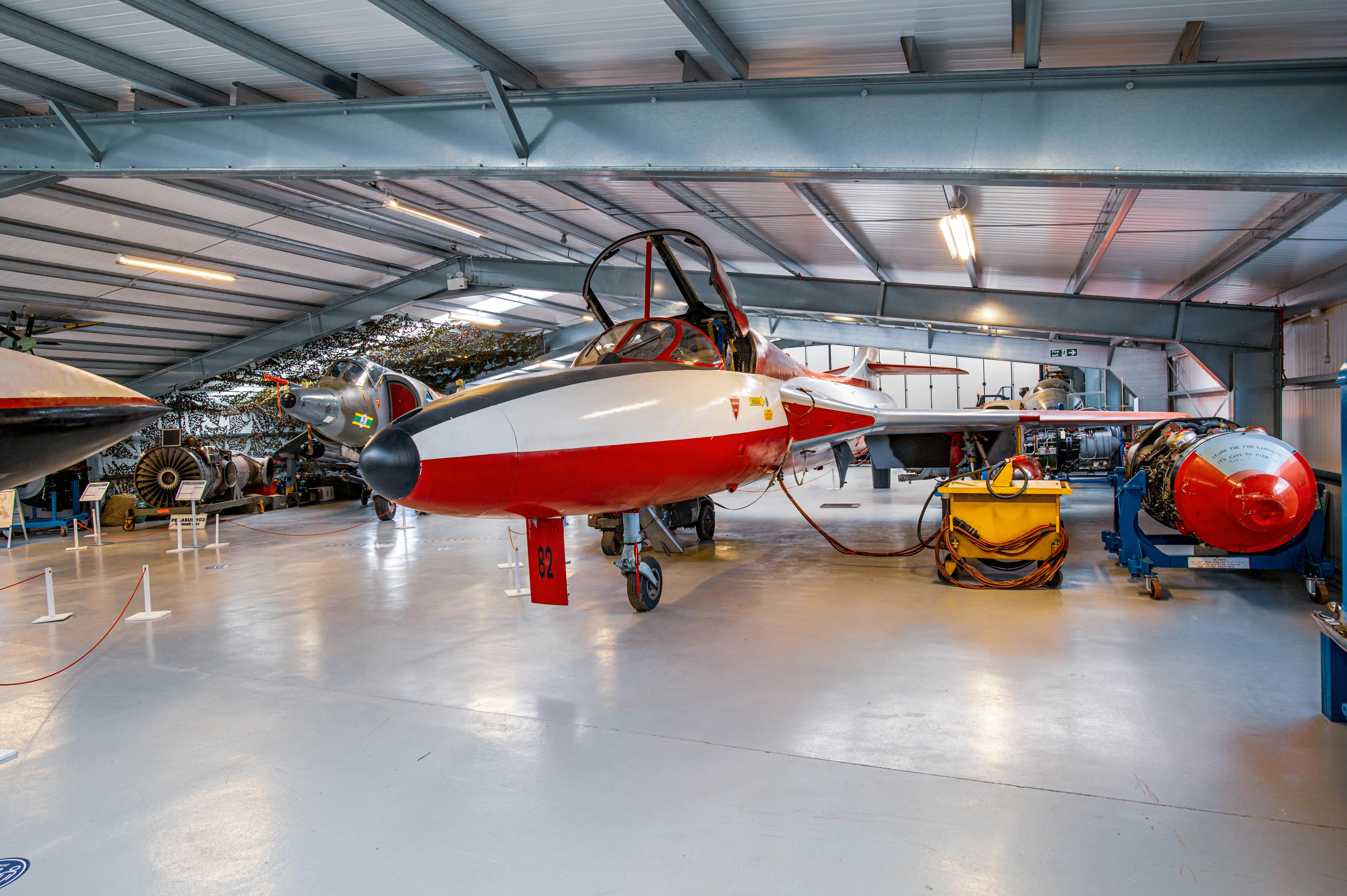 Interior view of a museum hangar displaying several vintage jet aircraft. A red and white jet with number 82 is in the foreground, with other aircraft and an engine on a stand in the background.