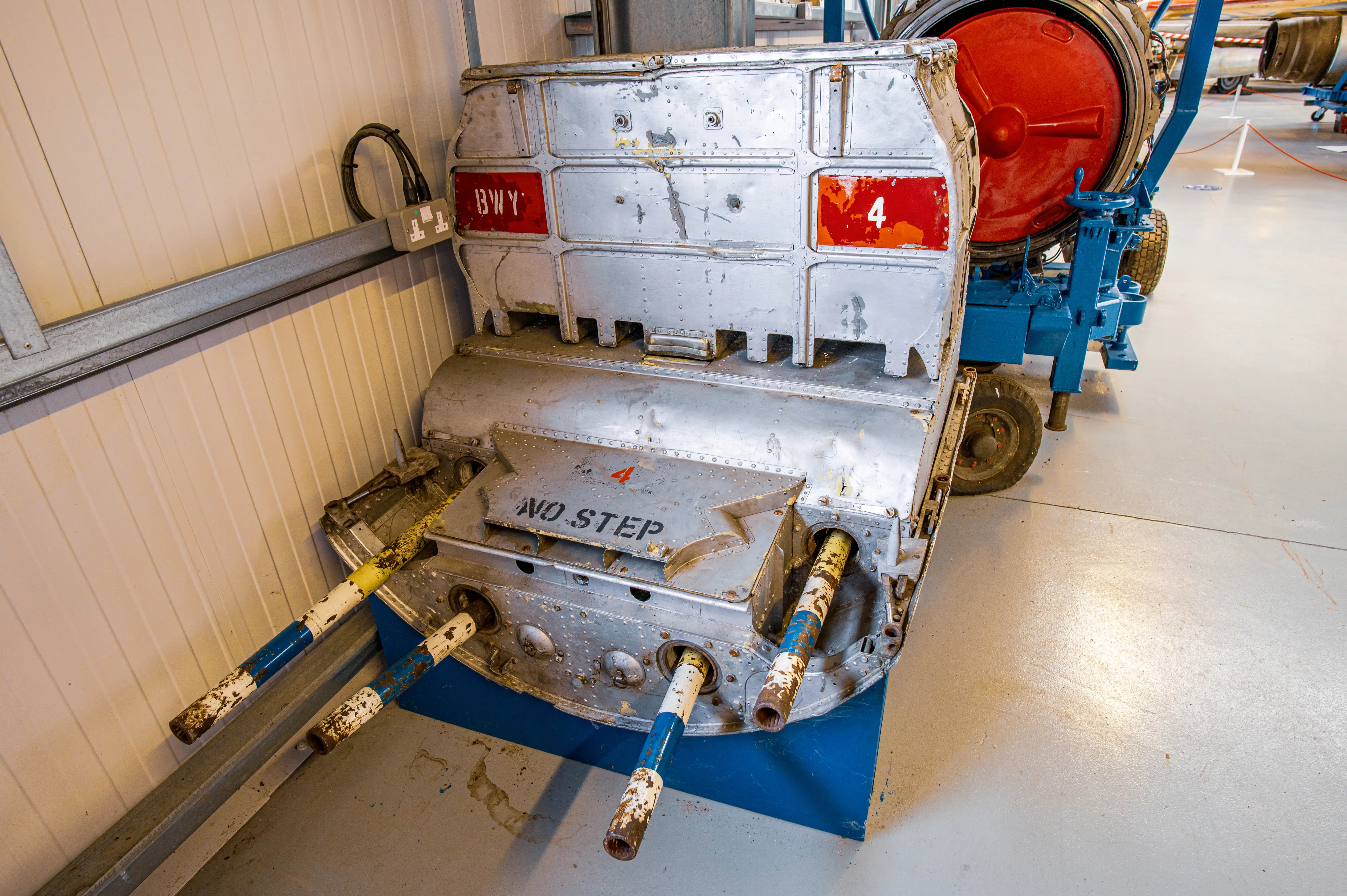 A metallic aircraft component with visible markings BWY and 4, and NO STEP, featuring multiple cylindrical tubes with blue and white bands, and a red circular object in the background.