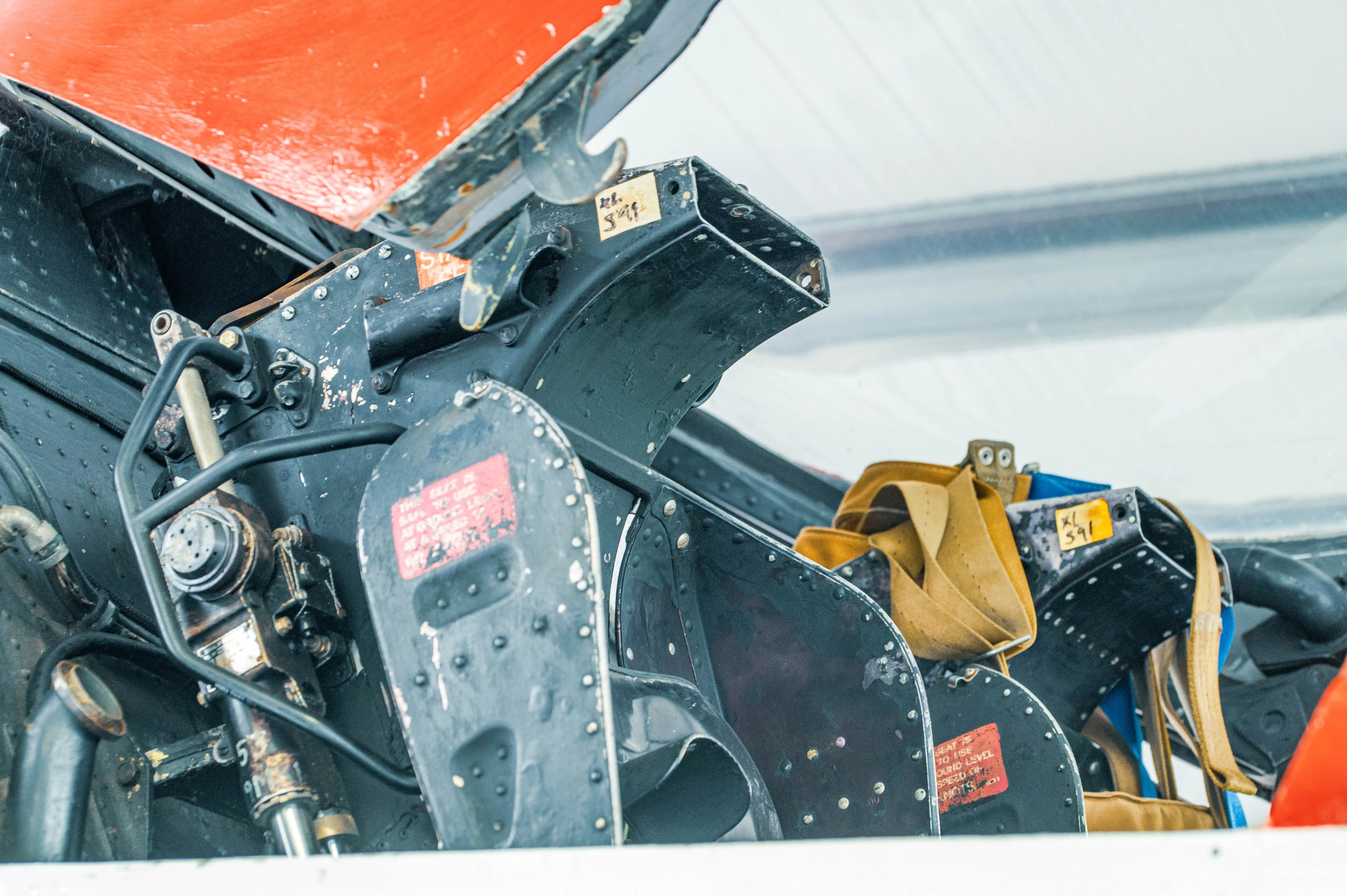 Detailed view of a vintage aircraft cockpit with black metal seats, tan harness straps, and orange canopy framing.