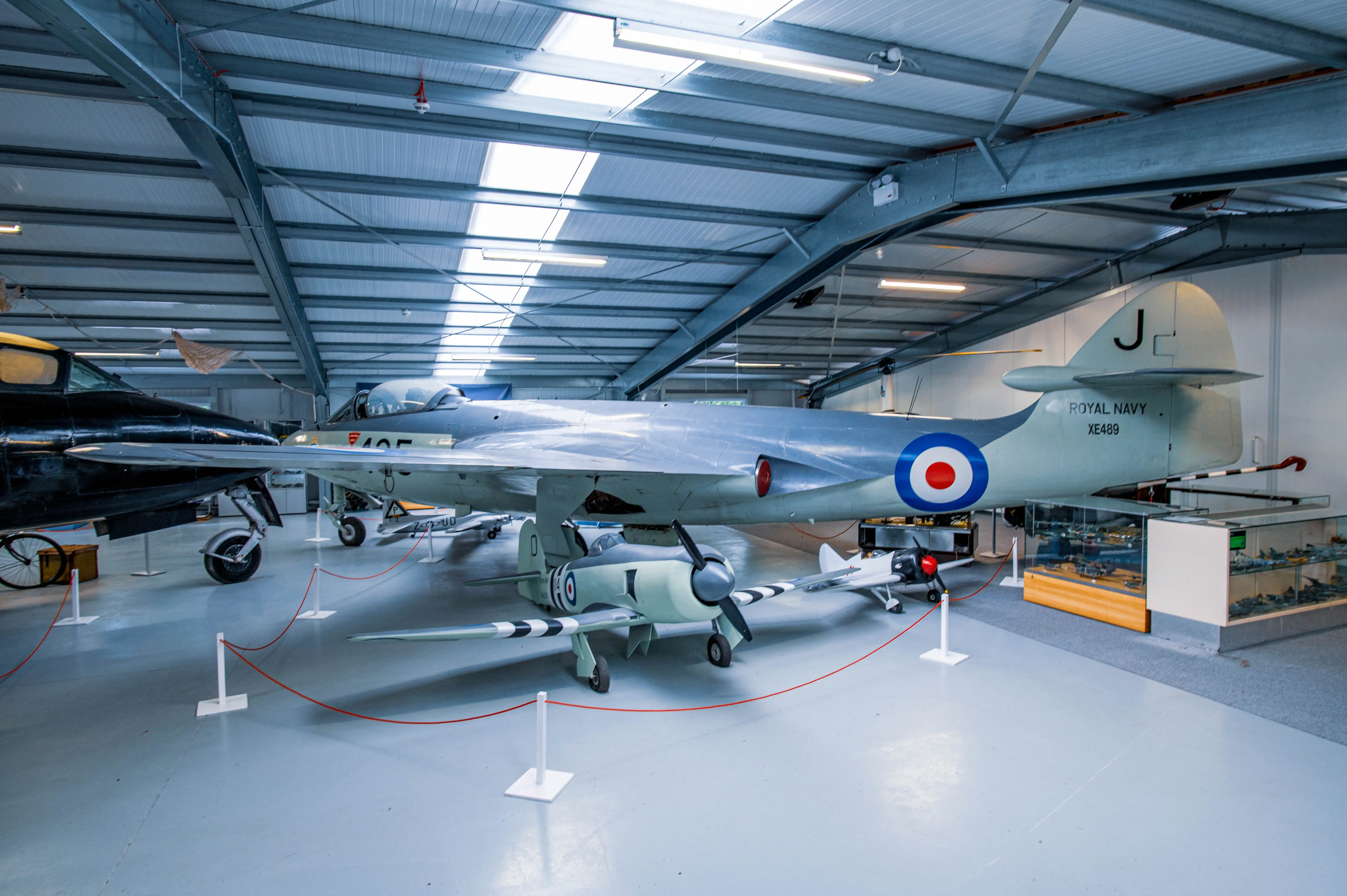 A de Havilland Sea Vixen jet aircraft, painted light grey with a Royal Navy roundel, is parked inside a hangar. In front of it sits a smaller, light green propeller aircraft. A black aircraft is partially visible to the left.
