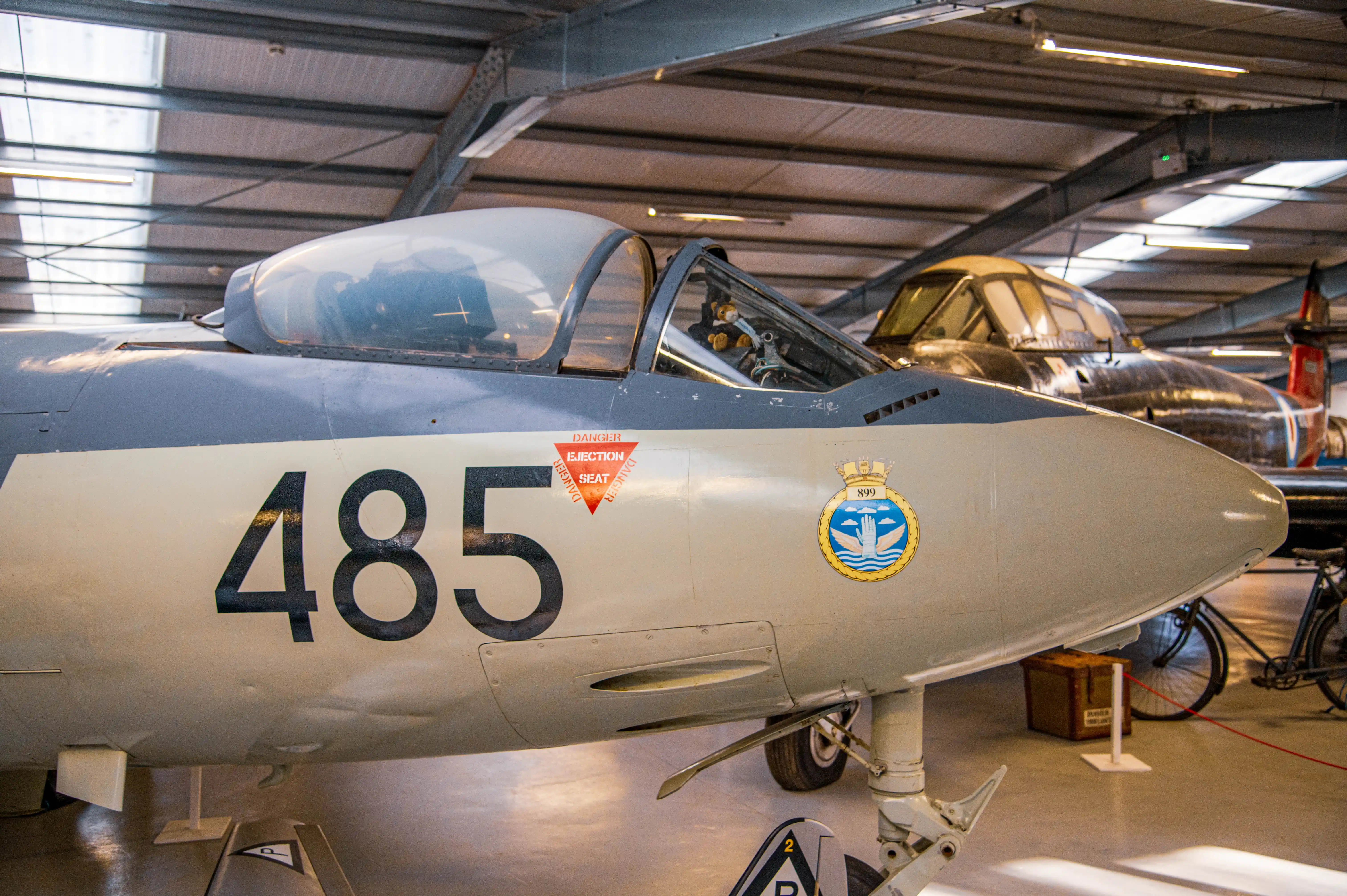 A side view of the cockpit and nose of a grey and cream coloured vintage jet fighter aircraft. The number 485 is painted in black on the cream section of the fuselage. A red triangle with the words DANGER EJECTION SEAT is visible to the right of the number. A circular crest with the number 899 is also present on the fuselage. In the background, another aircraft and a bicycle can be seen within a museum hangar.