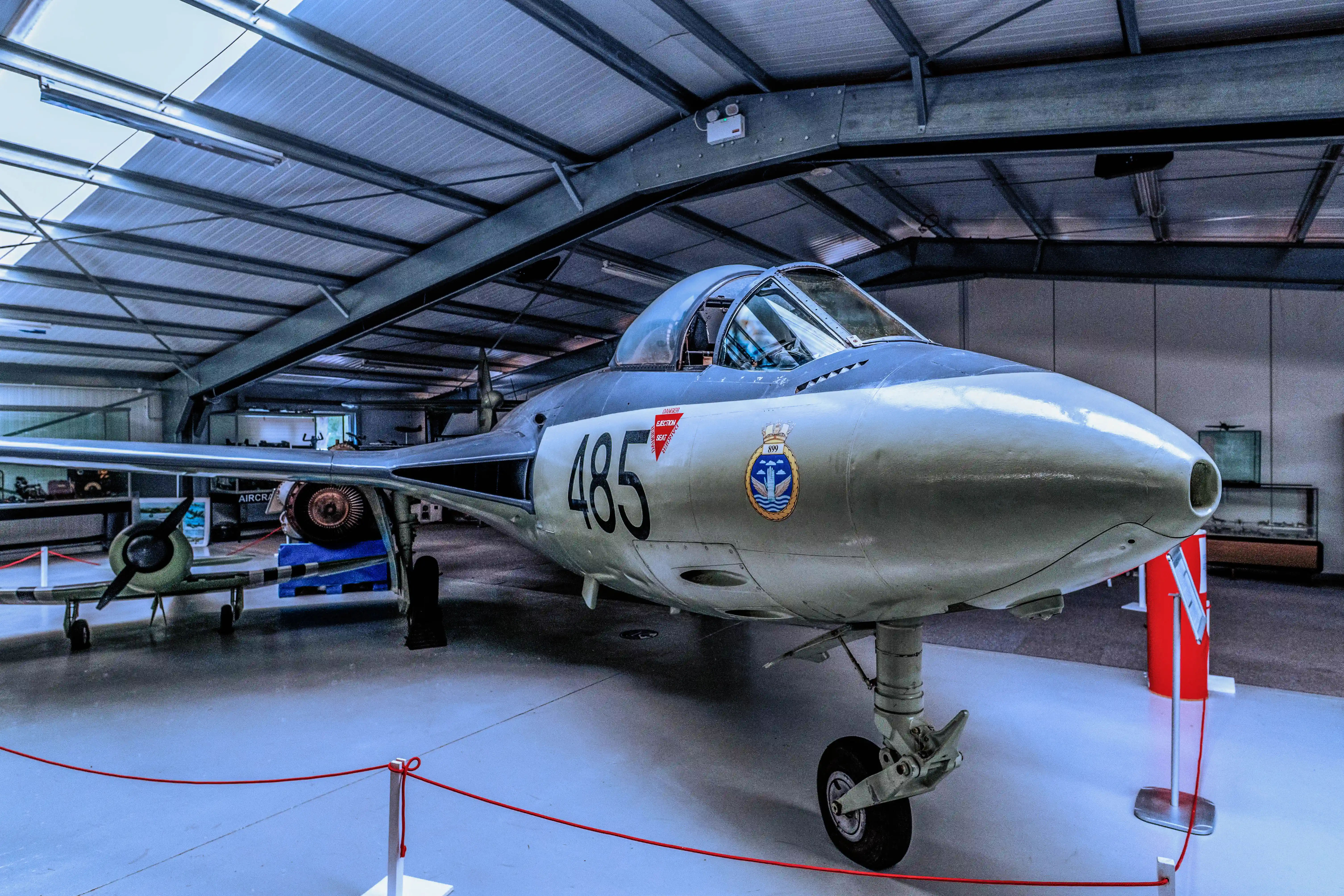 A striking view of a vintage Royal Navy jet fighter, bearing the number 485, showcased within a museum hangar. The aircraft's sleek design and prominent markings are clearly visible against the industrial backdrop.