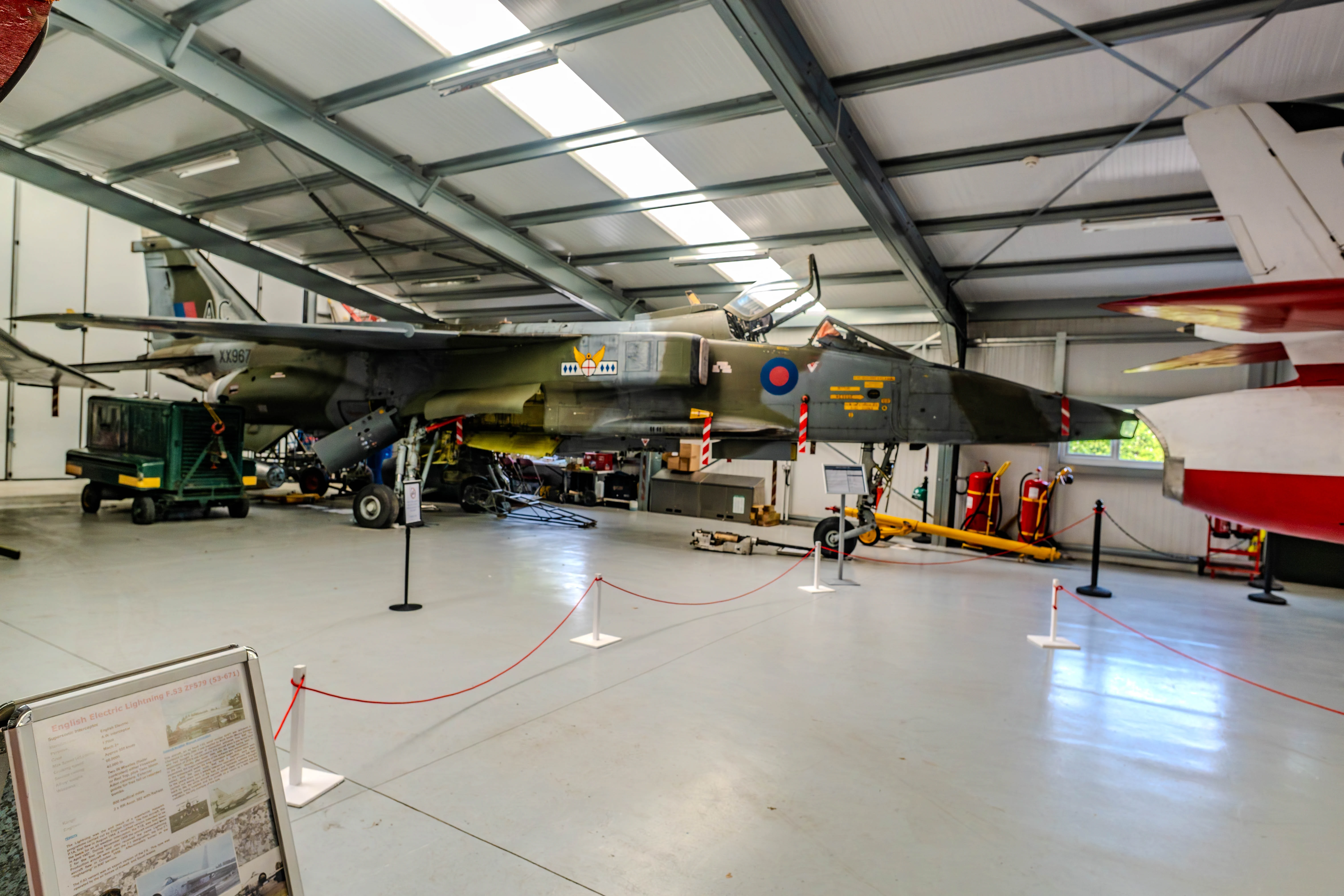 A British military jet fighter, the SEPECAT Jagua (XX967), is parked indoors in a museum hangar. The aircraft is painted in a camouflage scheme and features its original markings and a red and white striped tail fin.