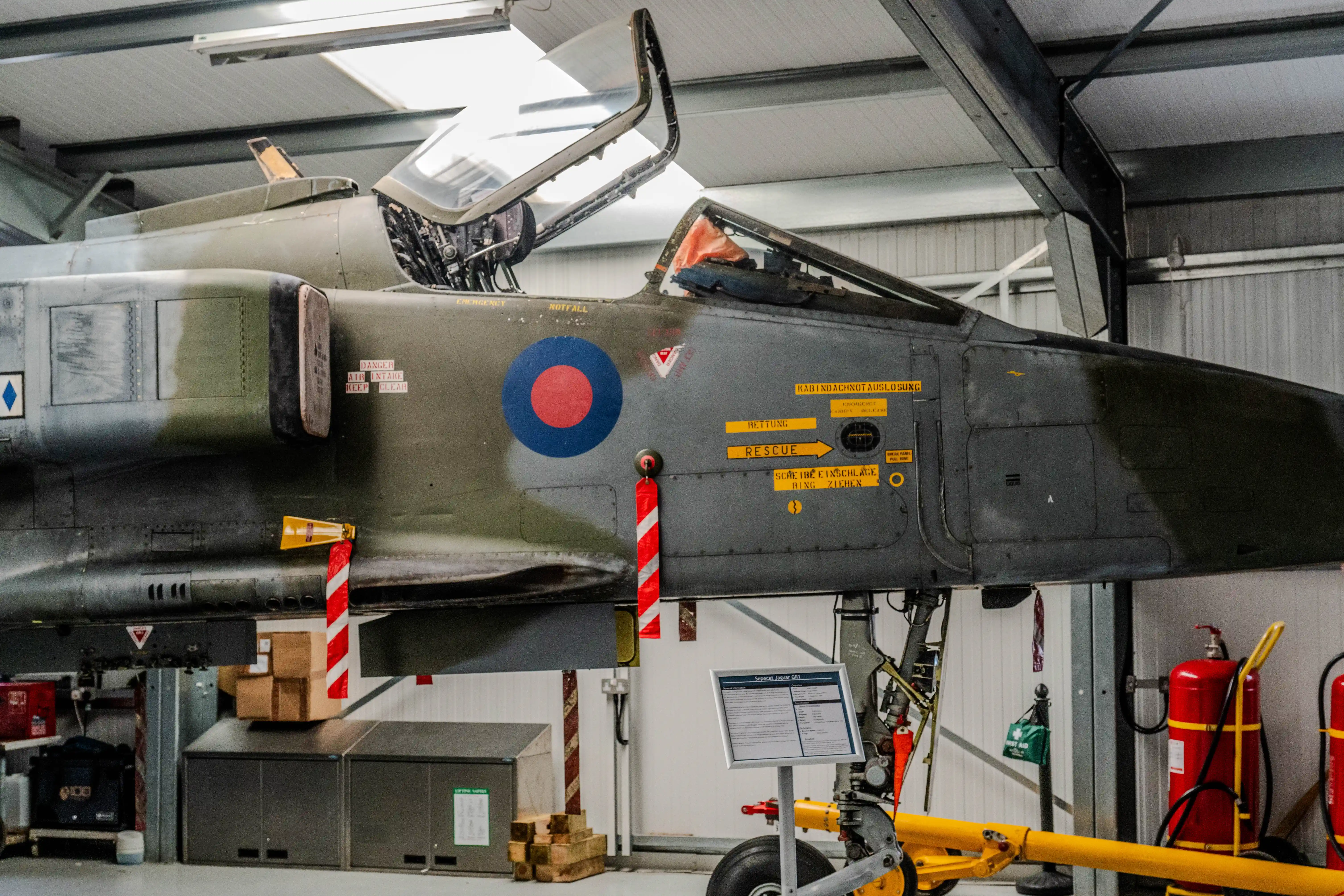 Close-up of a vintage jet fighter aircraft in a hangar, showing the cockpit area, national insignia, and various warning labels.