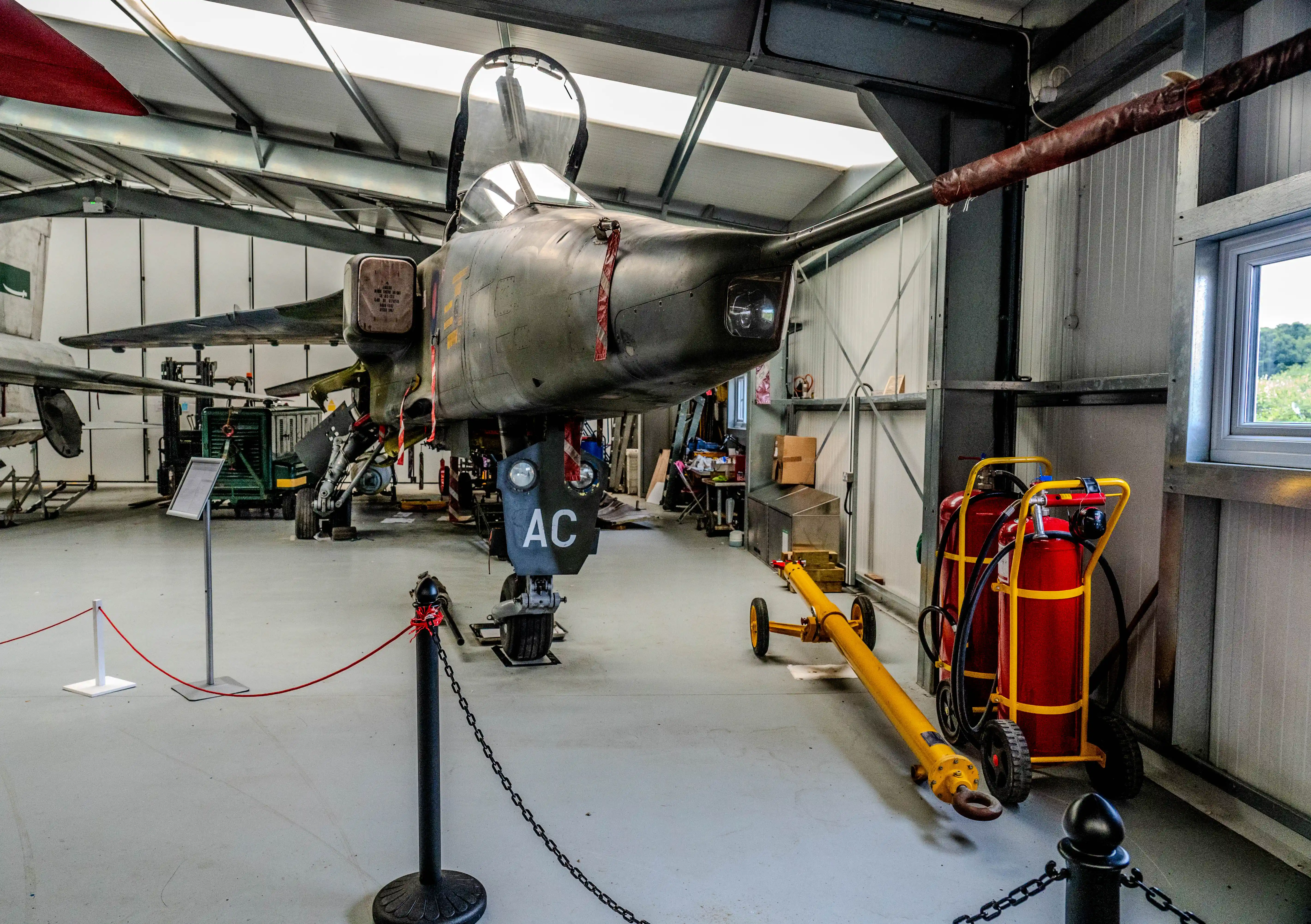 A close-up view of the front of a dark-coloured jet fighter plane, resting on its landing gear inside a modern industrial hangar. Yellow and red fire extinguishers are positioned to the right of the aircraft, along with a yellow tow bar.