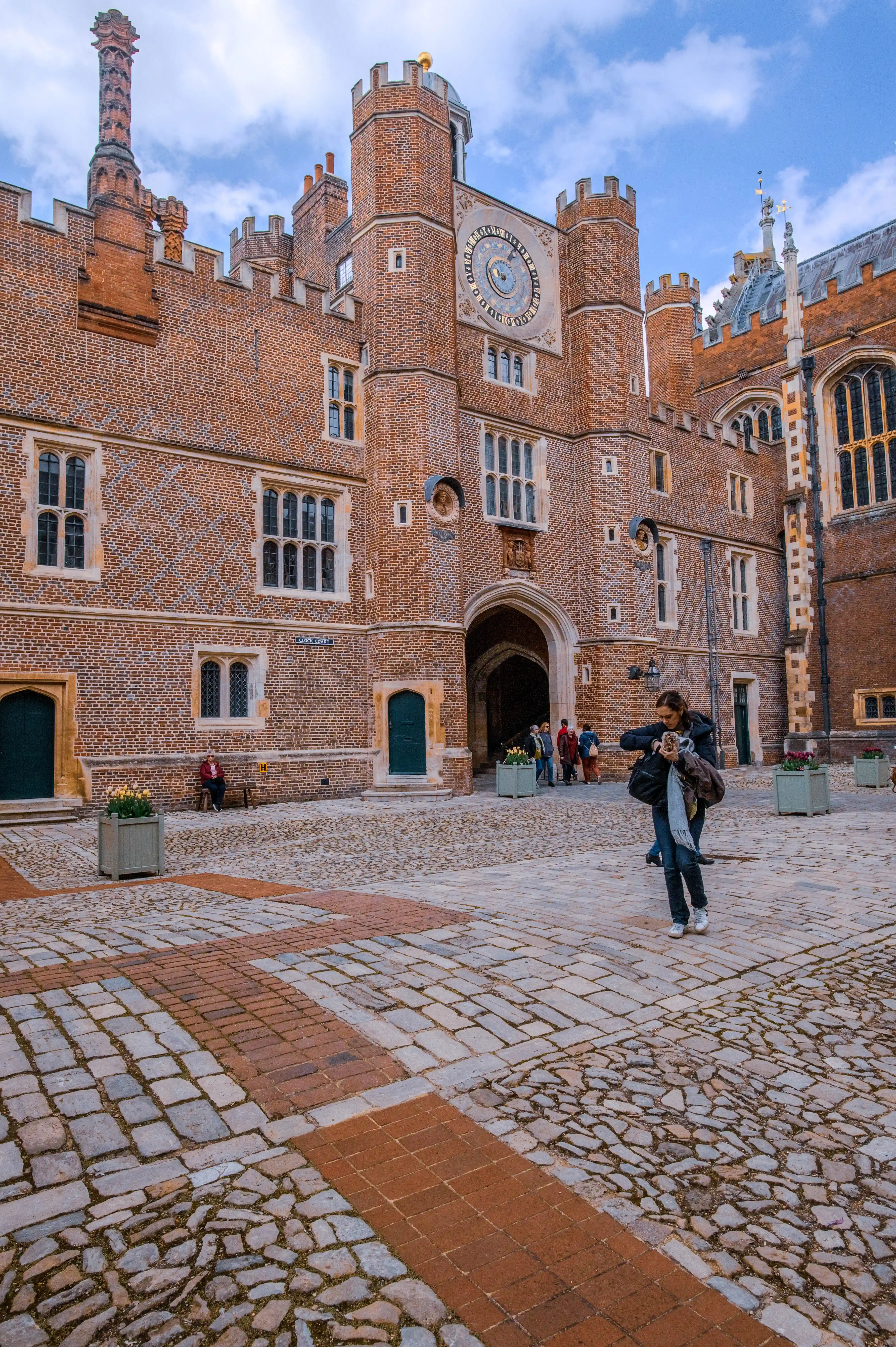 A cobbled courtyard at Hampton Court Palace, with the Great Clock Tower featuring a large clock face in the background. Several people are visible in the courtyard, with one person taking a photograph.