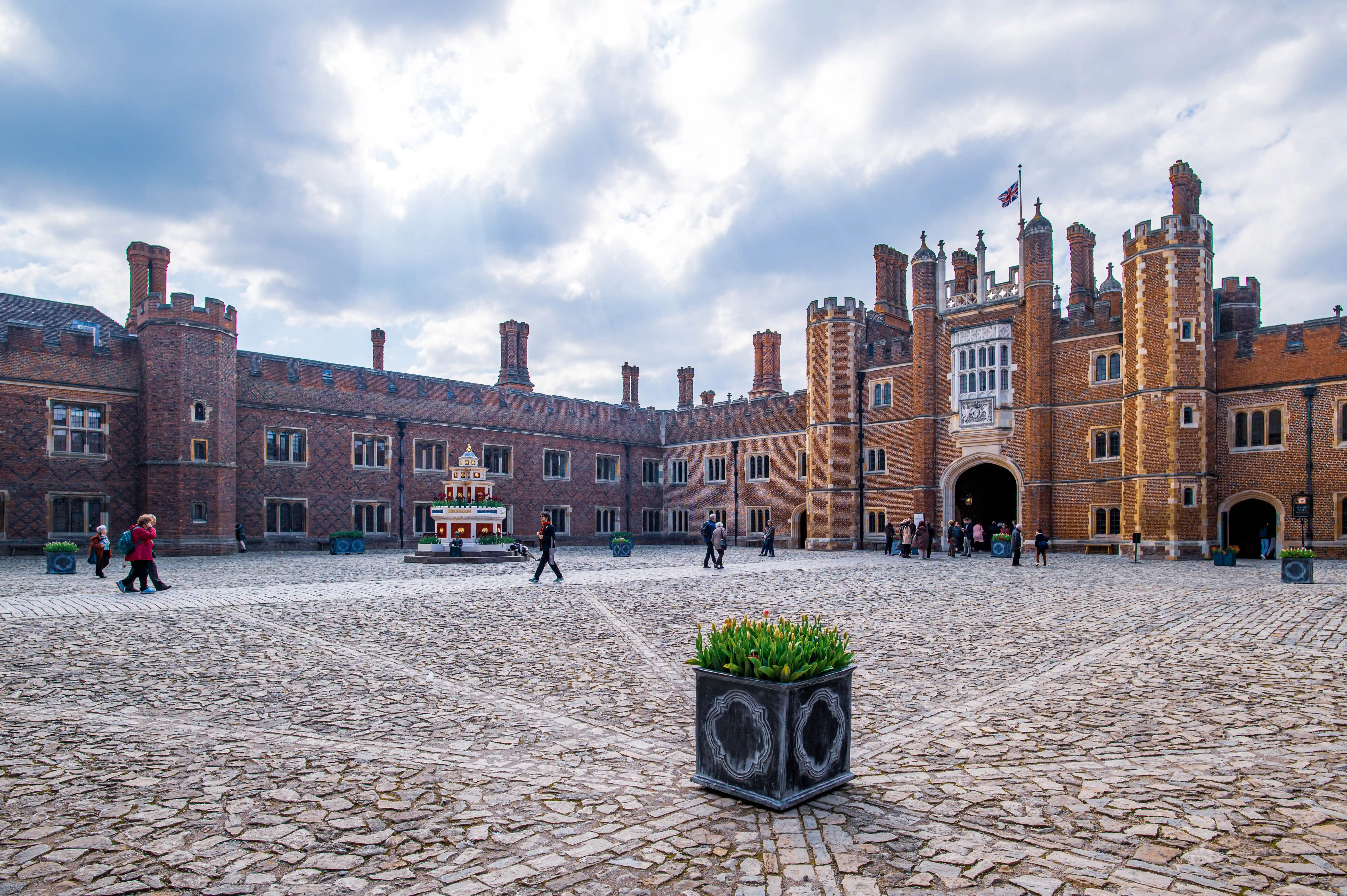 Wide shot of the cobbled courtyard at Hampton Court Palace. A grand red-brick Tudor building with many chimneys forms the backdrop. A large arched entrance is visible, with people gathered around. The Union Jack flag flies overhead. In the foreground, a stone planter holds a bunch of tulips.