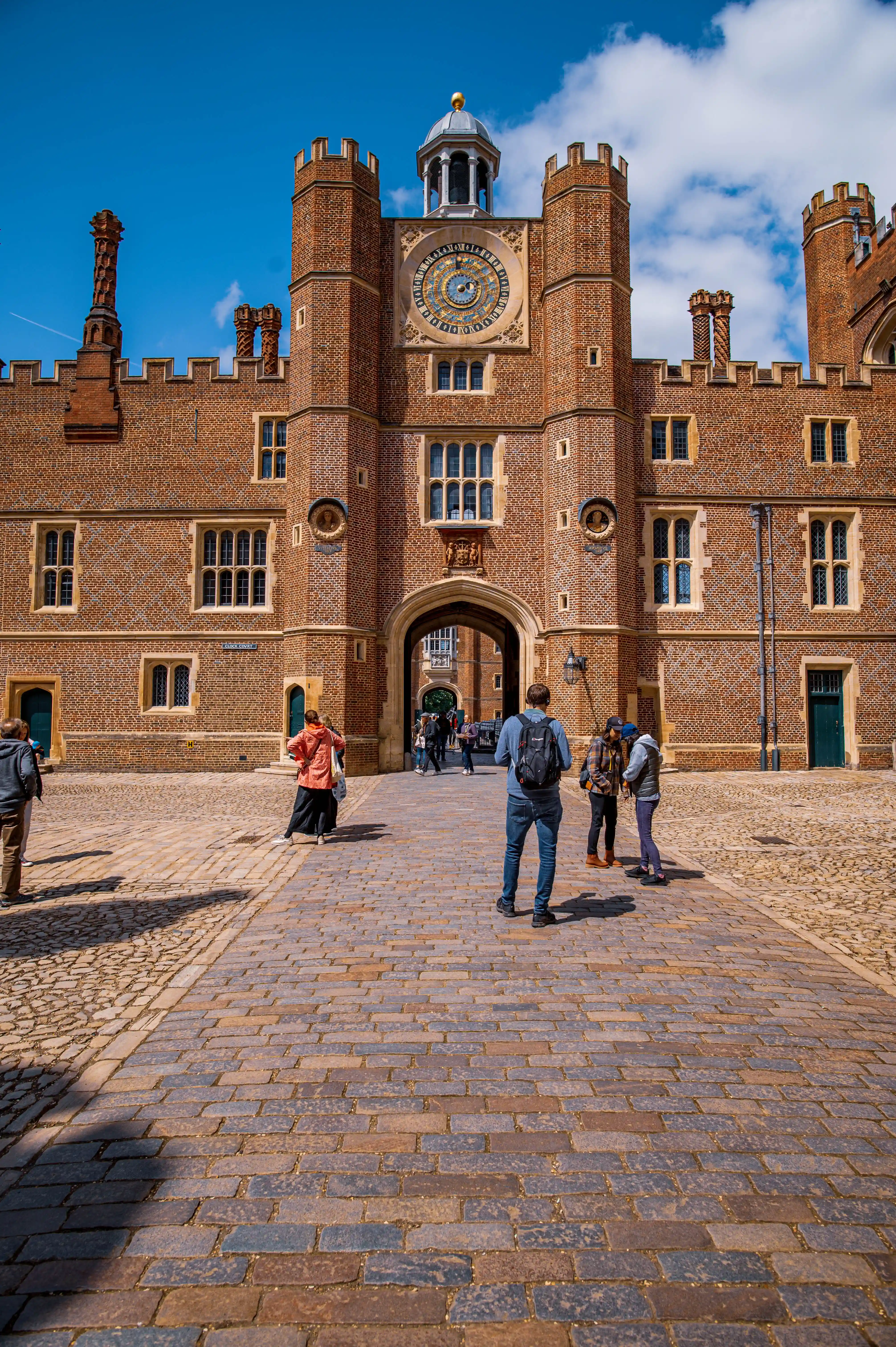 The Clock Tower of Hampton Court Palace stands tall against a blue sky with scattered clouds. The brick facade of the historic building is adorned with ornate windows and battlements. A large clock face is prominently displayed on the tower. In the foreground, a cobblestone courtyard is populated by a few tourists.