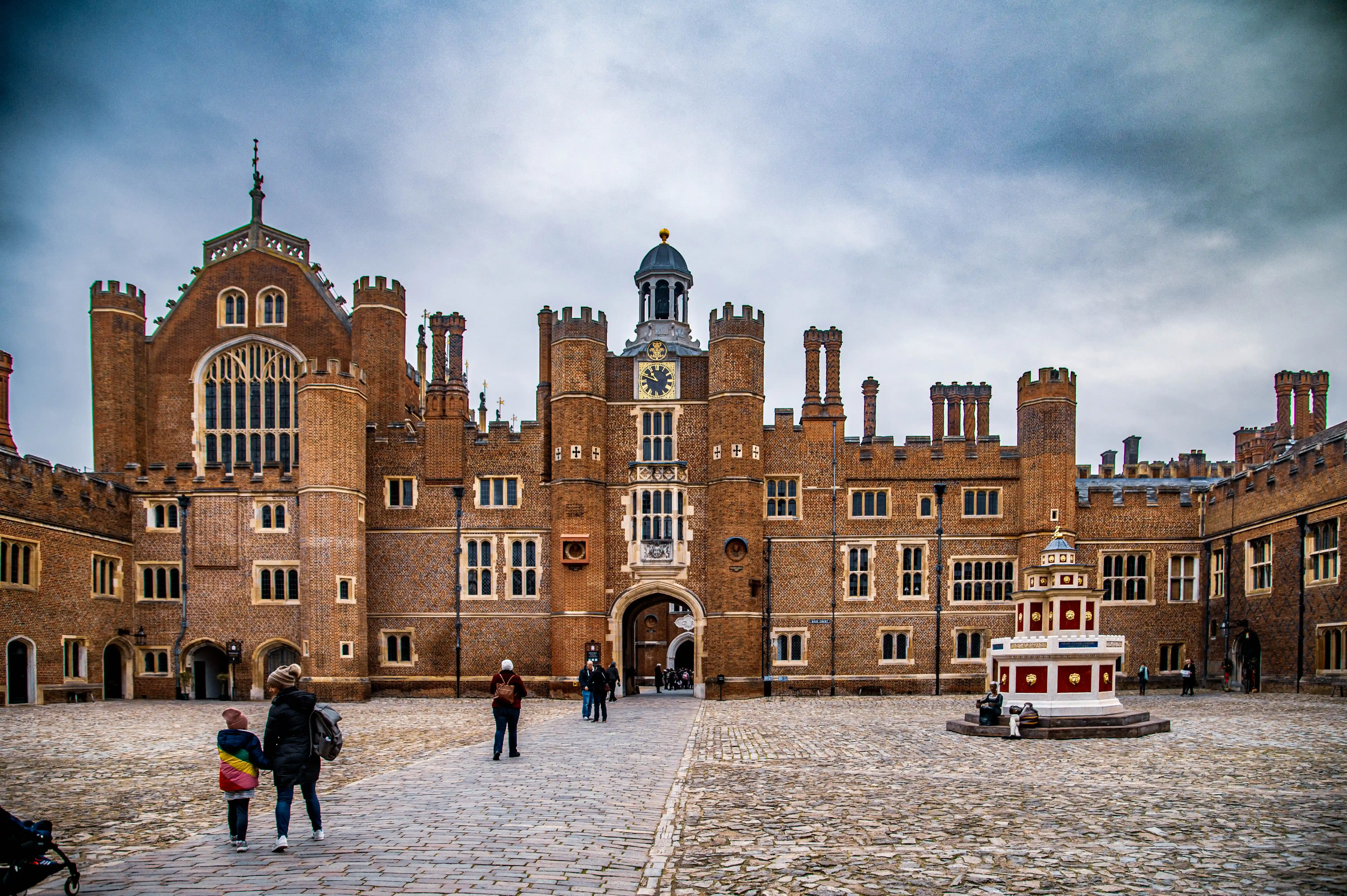Exterior view of Hampton Court Palace's courtyard, showcasing its brick facade, central clock tower, and a decorative fountain. Two people are walking in the courtyard.