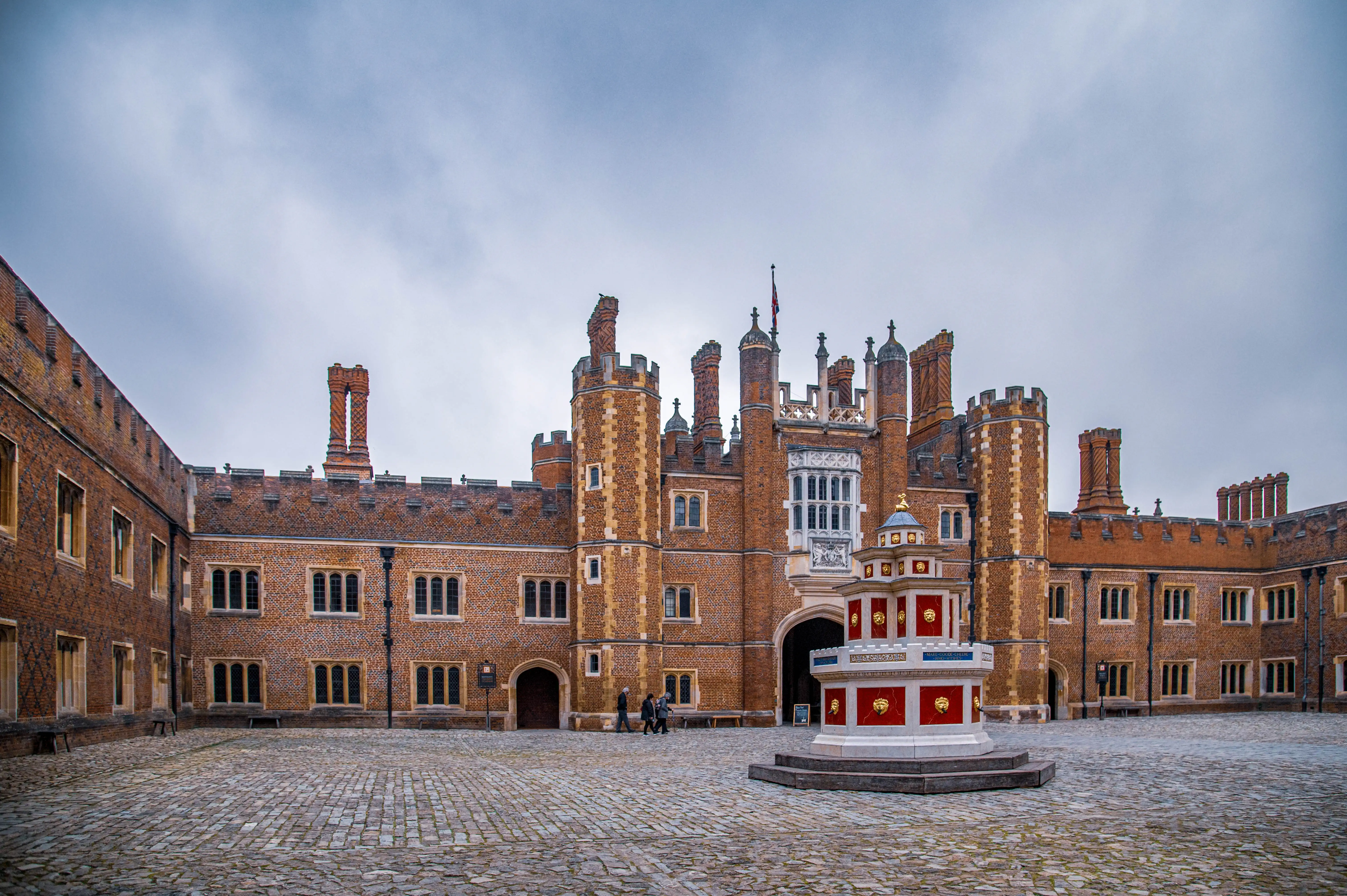A wide shot of the inner courtyard of Hampton Court Palace, showcasing its red brick Tudor architecture with tall chimneys and ornate windows. In the centre of the cobblestone courtyard is a red and white decorative fountain.