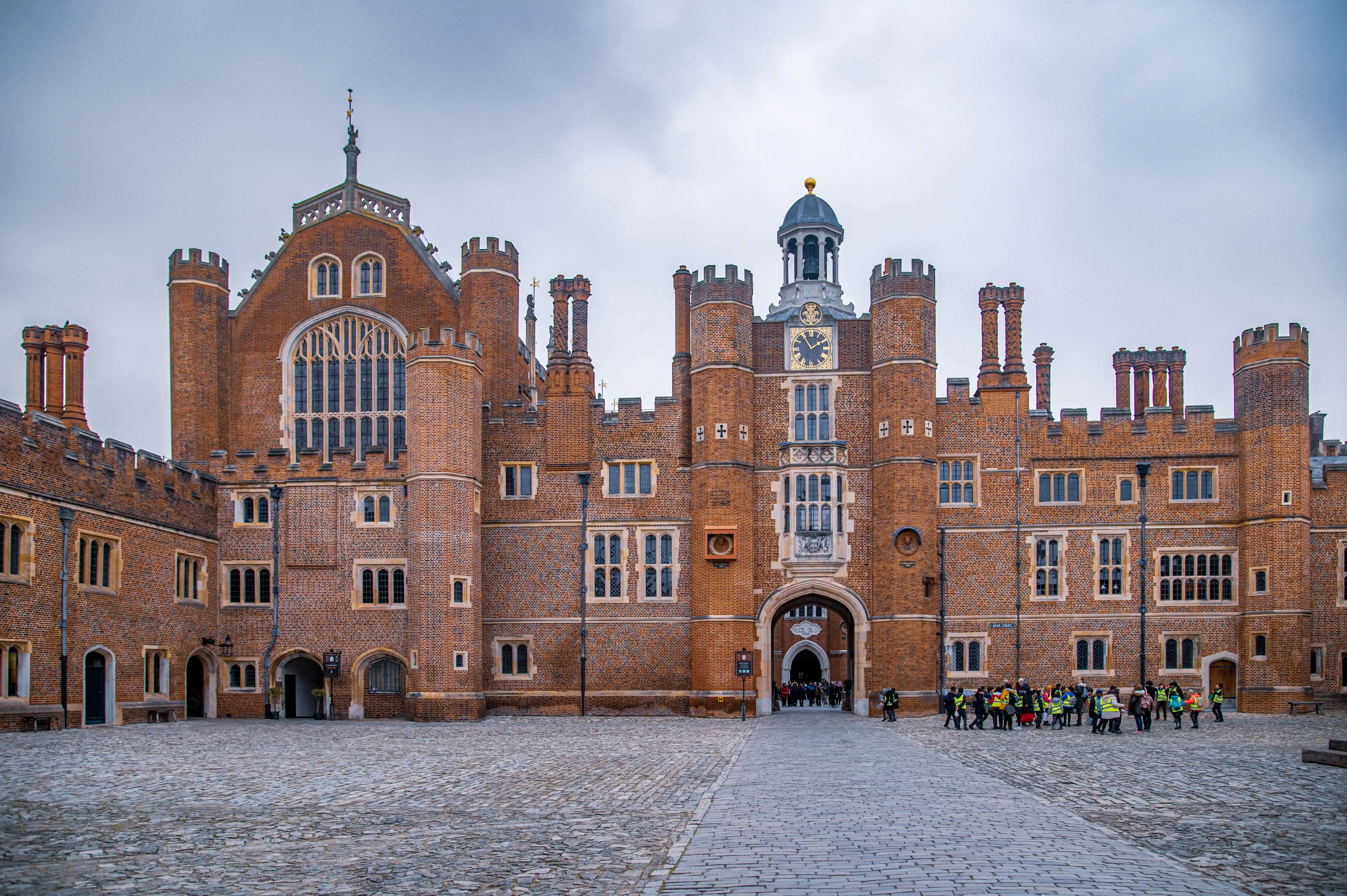 A wide shot of the courtyard of Hampton Court Palace, showing its distinctive red brick Tudor architecture, including towers, arched gateways, and many windows. A group of people are visible in the distance on the right side of the courtyard.