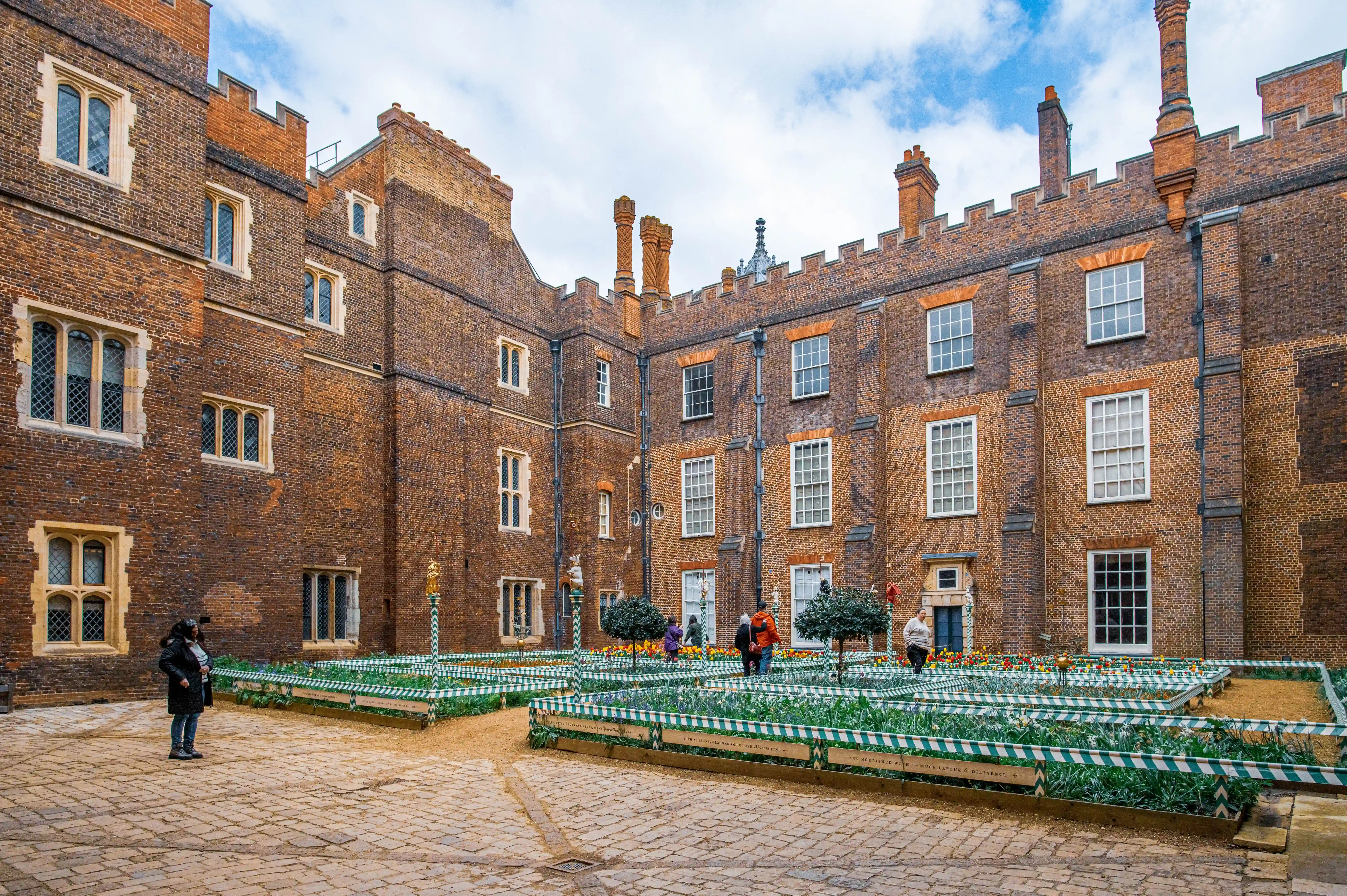 A wide shot of a historic brick courtyard at Hampton Court Palace, UK. In the centre is a formal garden with patterned flowerbeds and small trees. Cobblestones pave the ground and a few people are walking around.