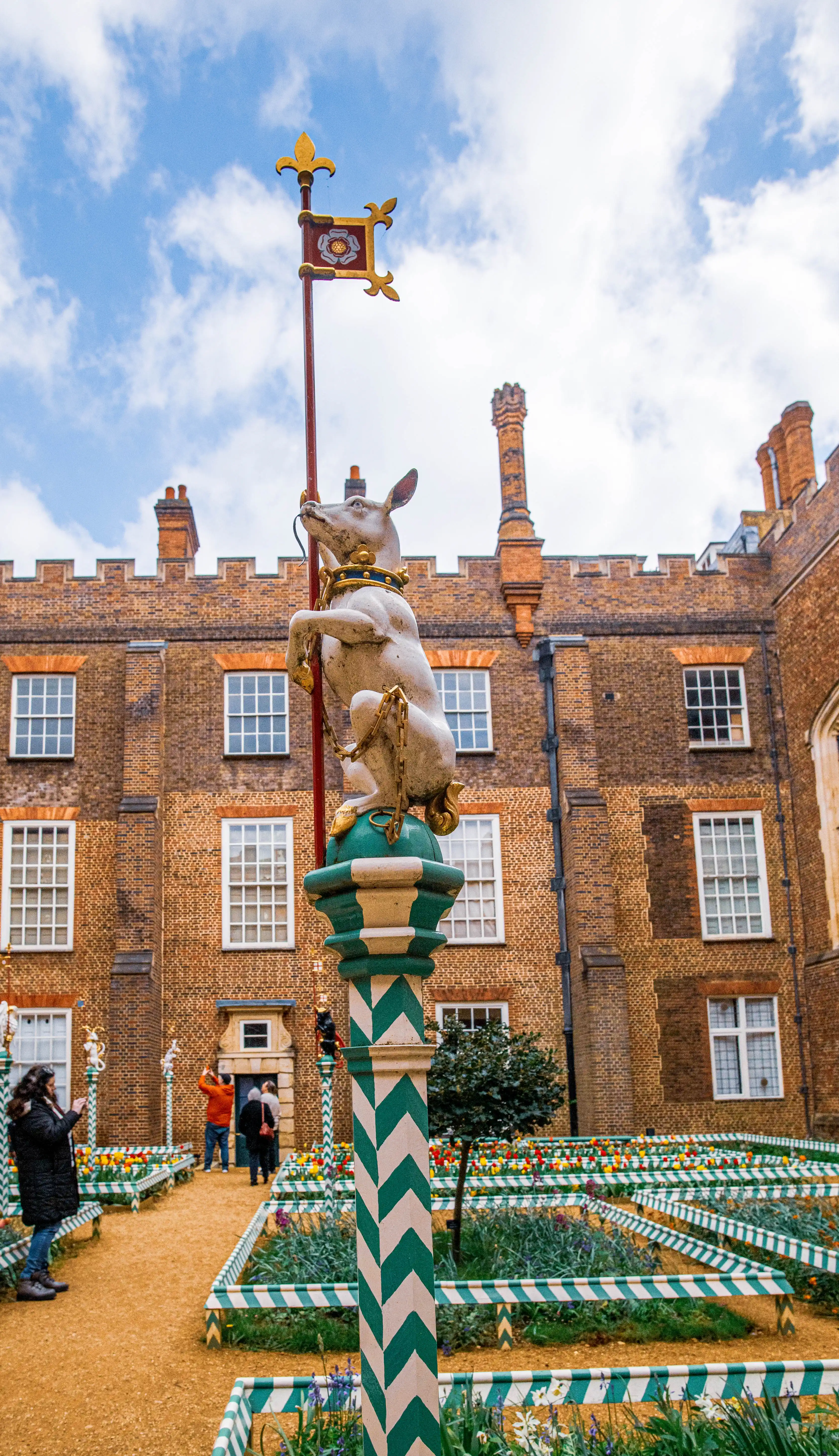 A white deer statue is positioned centrally in front of a red brick building. The statue is chained and stands on a green and white striped pedestal. A flagpole with a Tudor rose emblem is behind the statue. Visitors are visible in the background, looking at gardens and the building.