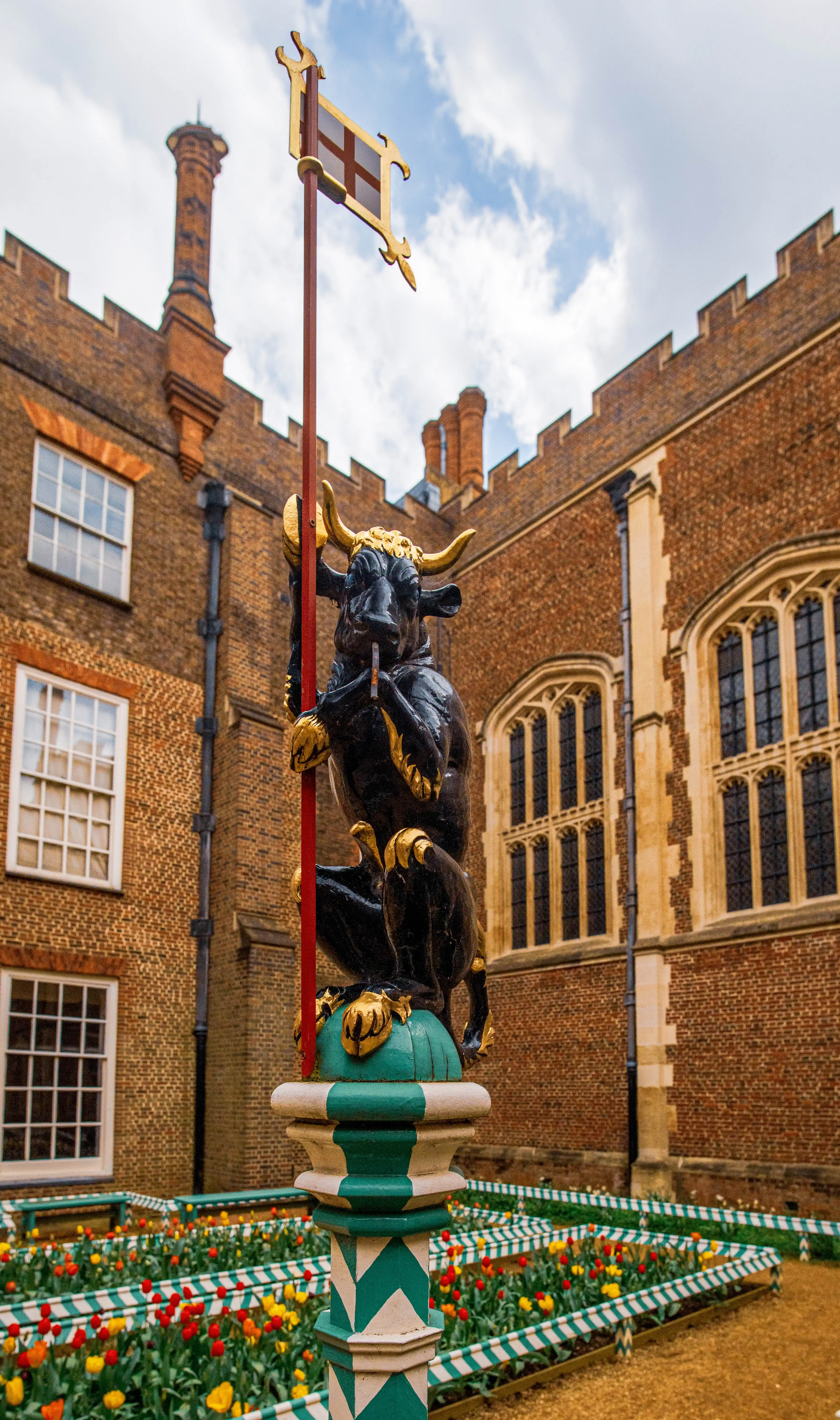 A black bull sculpture with golden horns and hooves, mounted on a decorative post, overlooks a garden filled with blooming tulips. The sculpture is in the foreground, with a red brick building and windows in the background.
