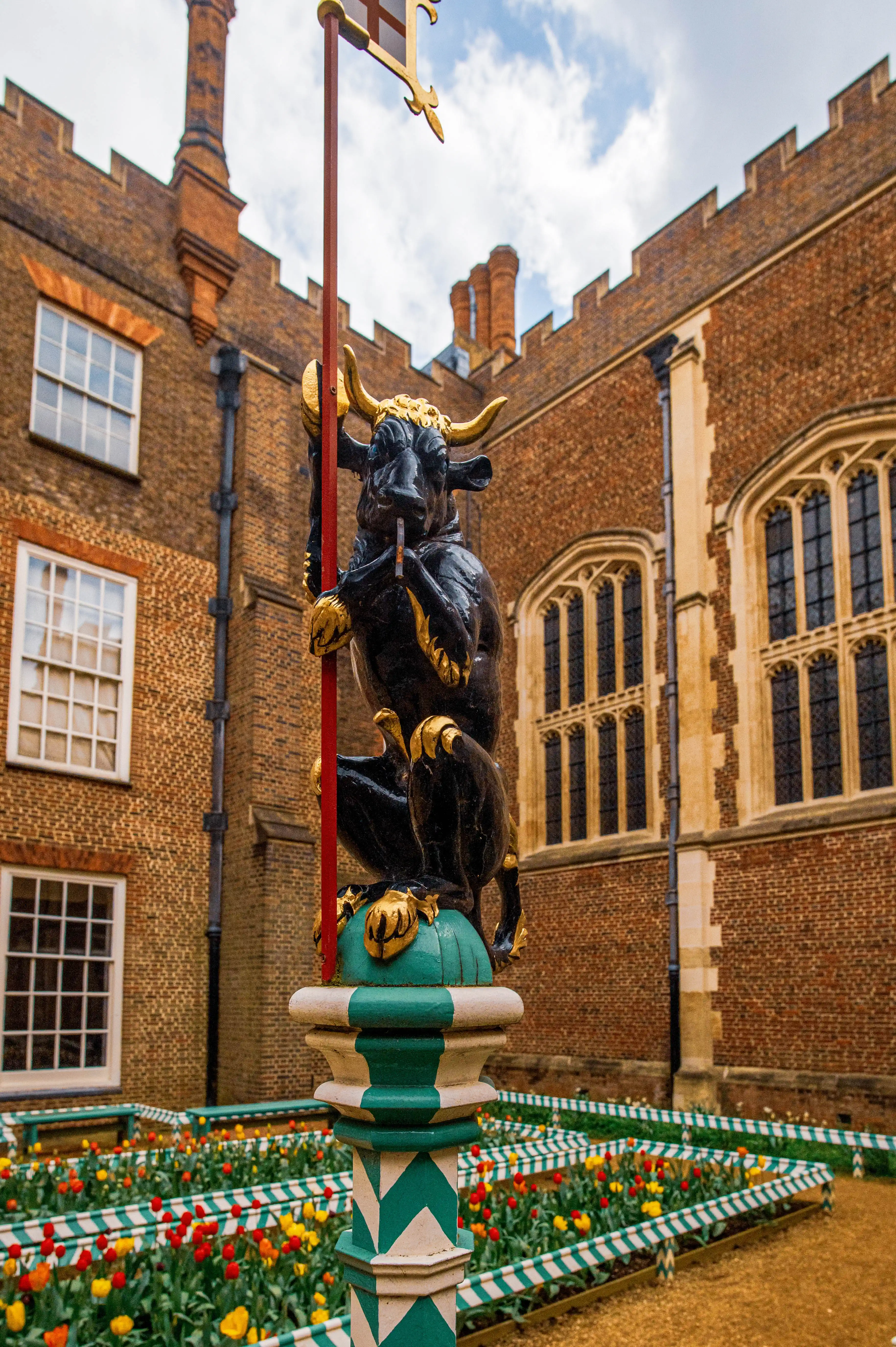 A black bull sculpture with golden horns and hooves, mounted on a decorative post, overlooks a garden filled with blooming tulips. The sculpture is in the foreground, with a red brick building and windows in the background.