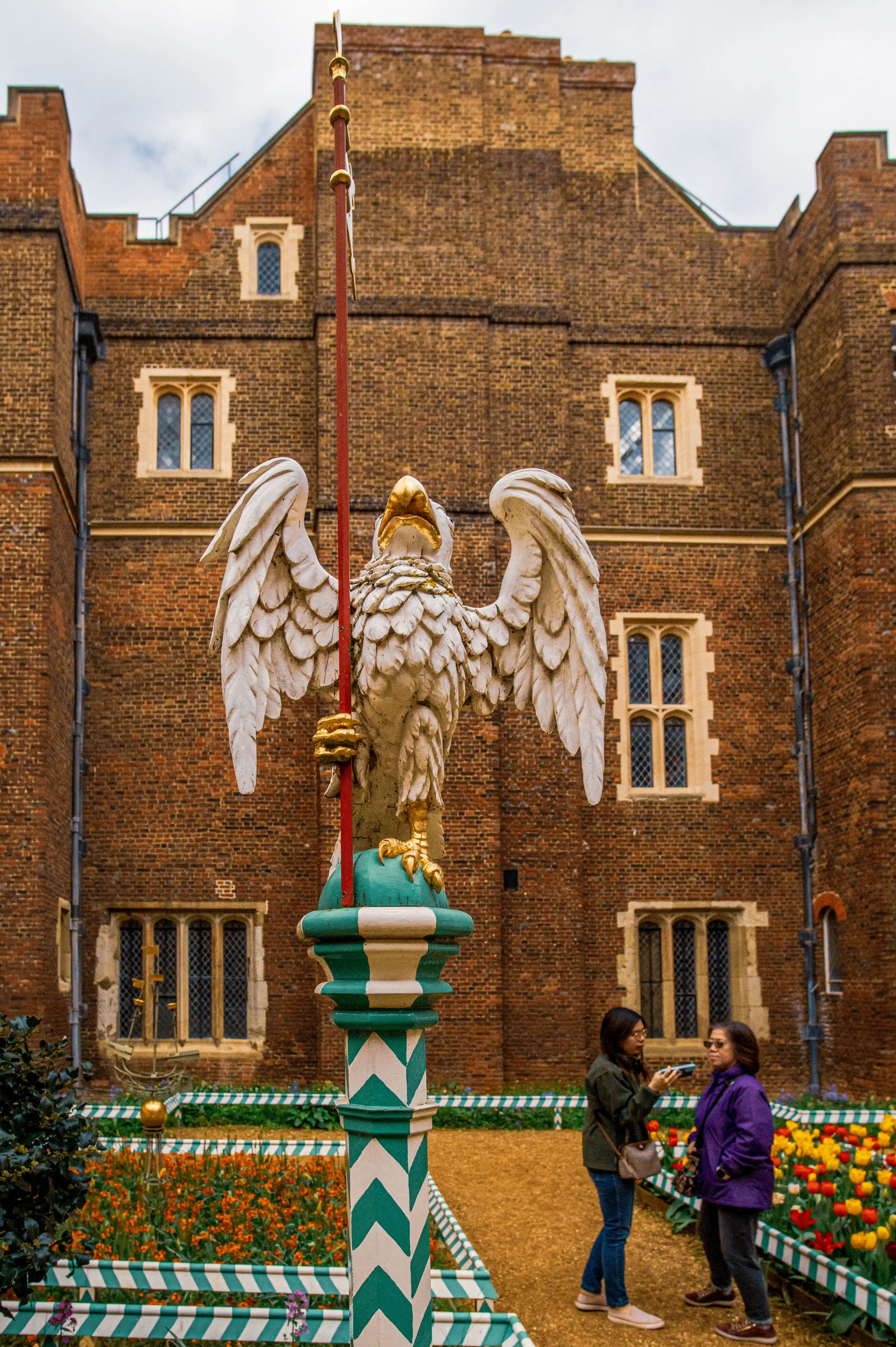 A stone statue of a white eagle with outstretched wings and a gold beak stands on a decorative pillar in front of a red brick building. Two women are standing to the right of the statue, looking at something in their hands. Flowerbeds with orange and yellow flowers are visible in the foreground and middle ground.