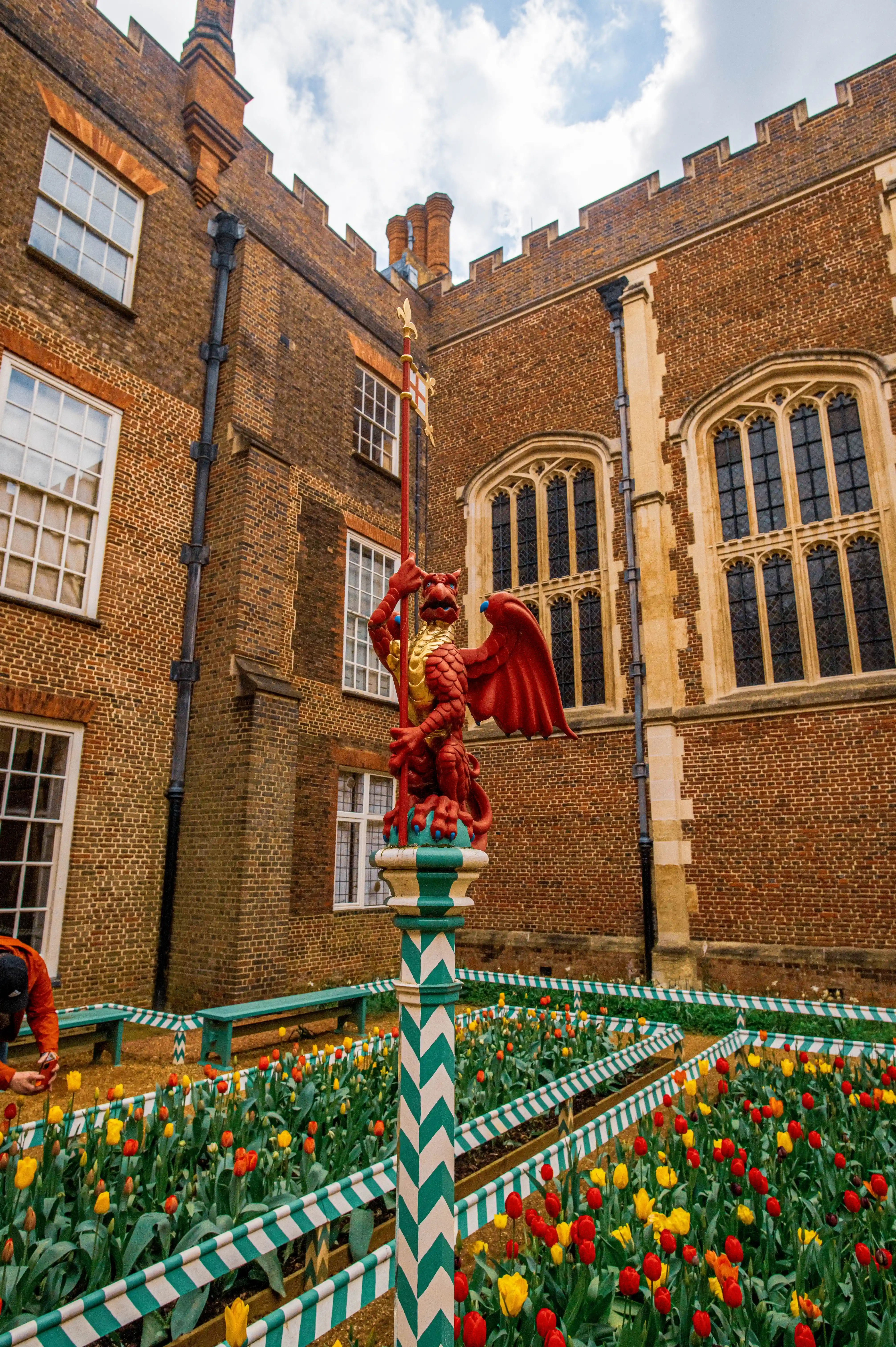 A red griffin statue stands in a courtyard filled with red and yellow tulips. The statue is on a patterned pillar and is surrounded by a flowerbed with green and white striped borders. Behind the garden are brick buildings with large windows.