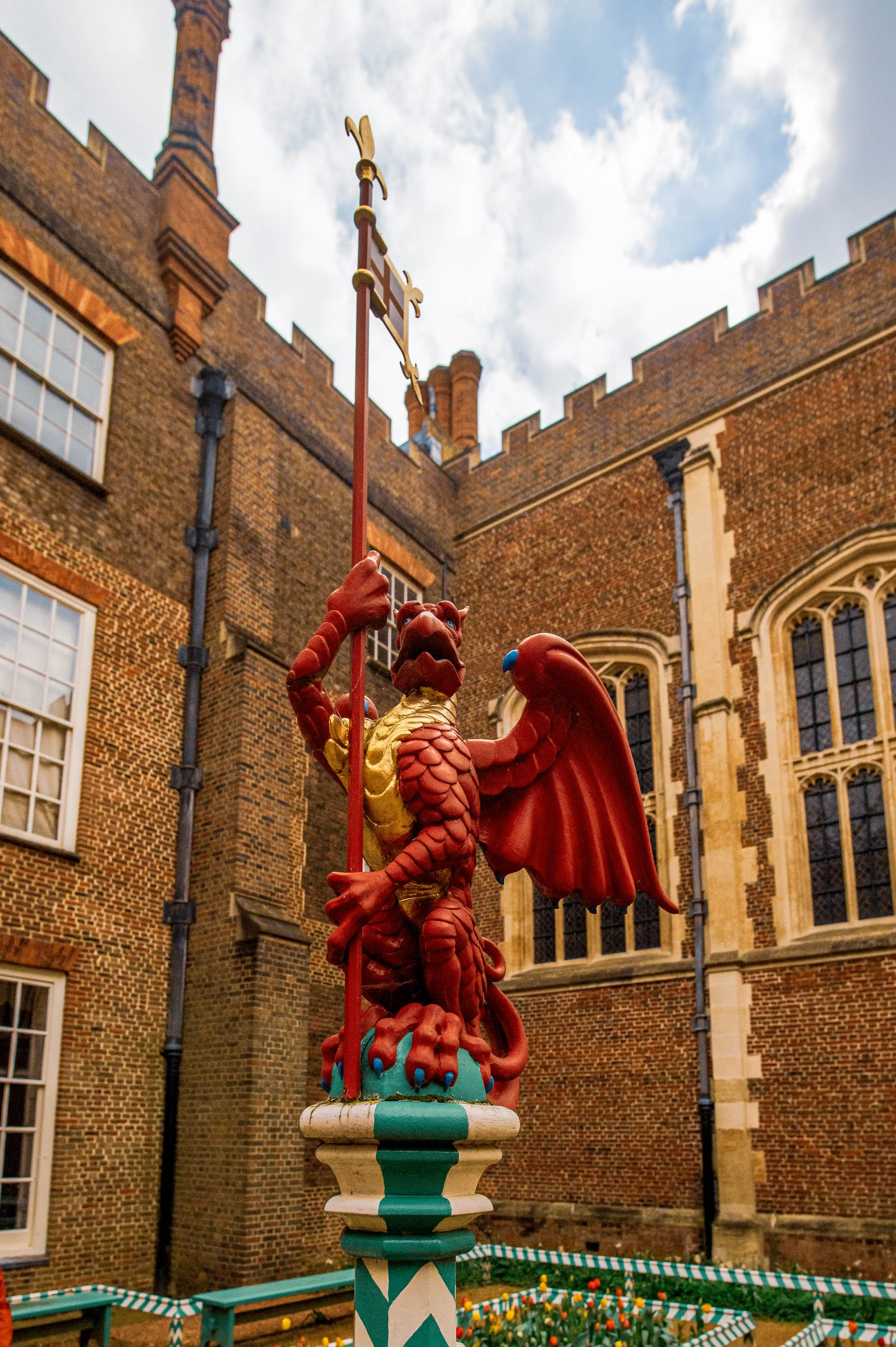 A red griffin statue stands in a courtyard filled with red and yellow tulips. The statue is on a patterned pillar and is surrounded by a flowerbed with green and white striped borders. Behind the garden are brick buildings with large windows.