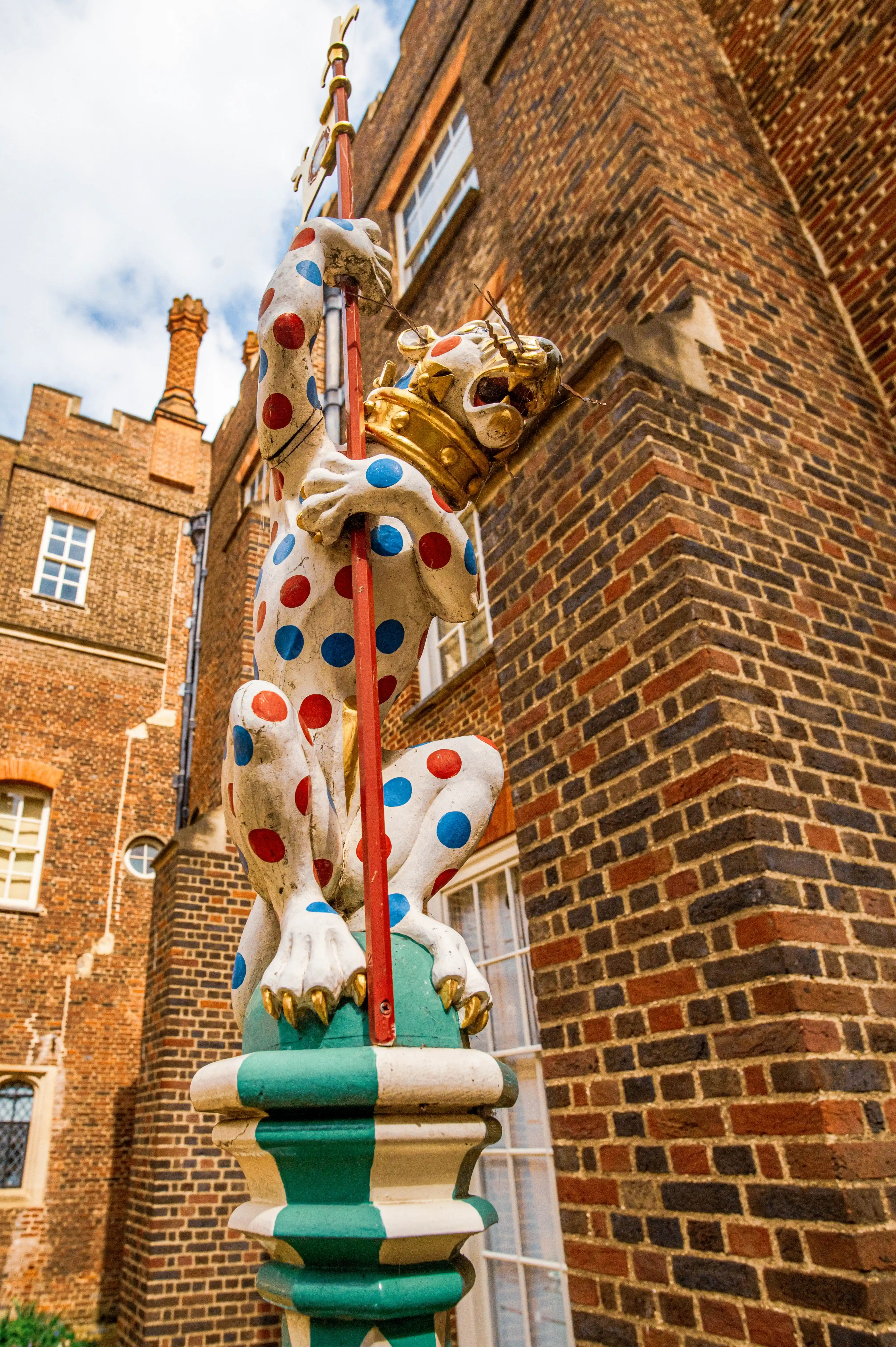 A low-angle shot of a fanciful statue of a crowned griffin covered in red and blue polka dots, holding a pole, with a brick building in the background.