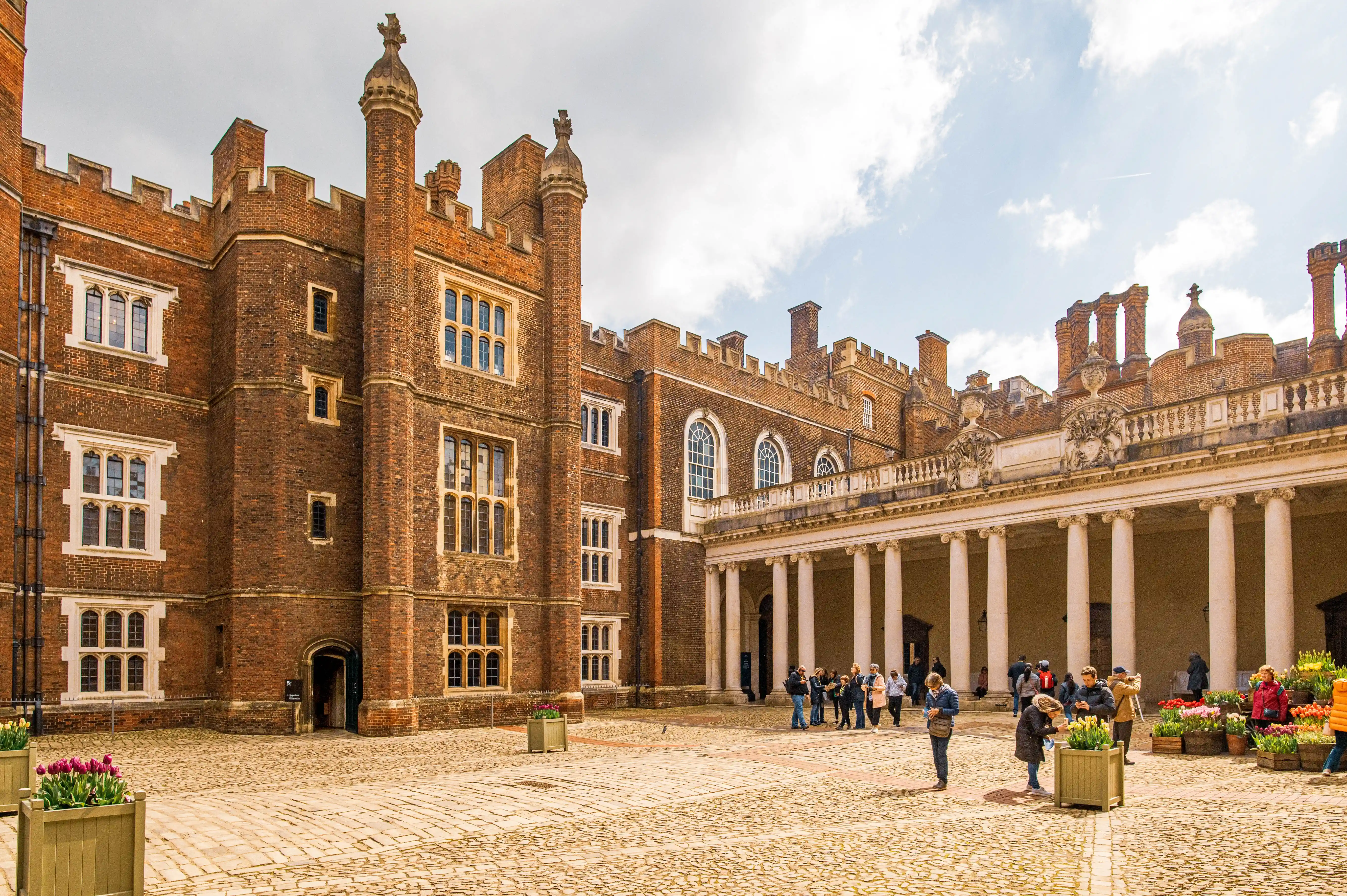 A wide shot of a courtyard at Hampton Court Palace, with people walking around, a stone-paved floor, and a building with a colonnade.