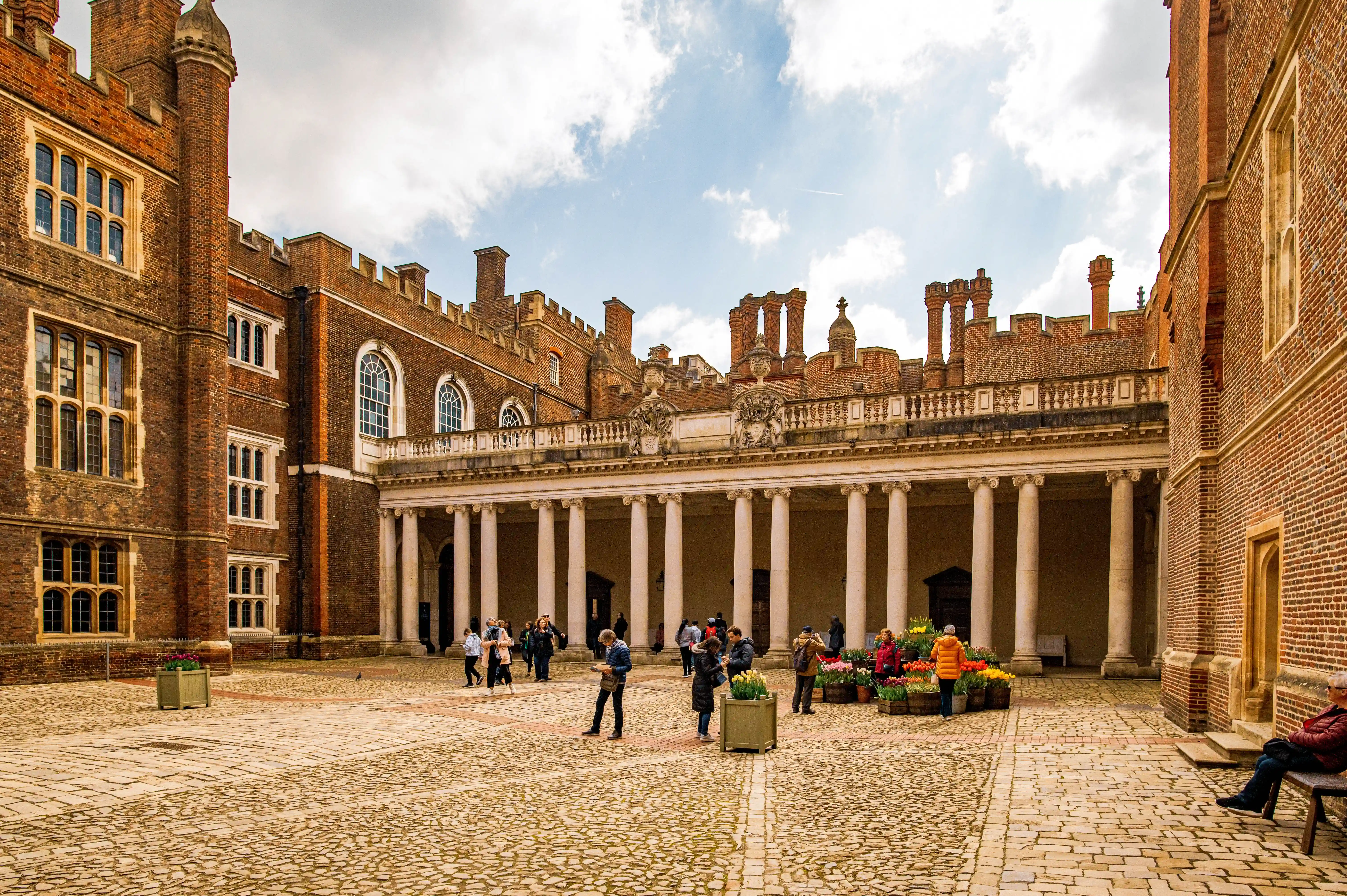 A wide shot of a courtyard at Hampton Court Palace, with people walking around, a stone-paved floor, and a building with a colonnade.