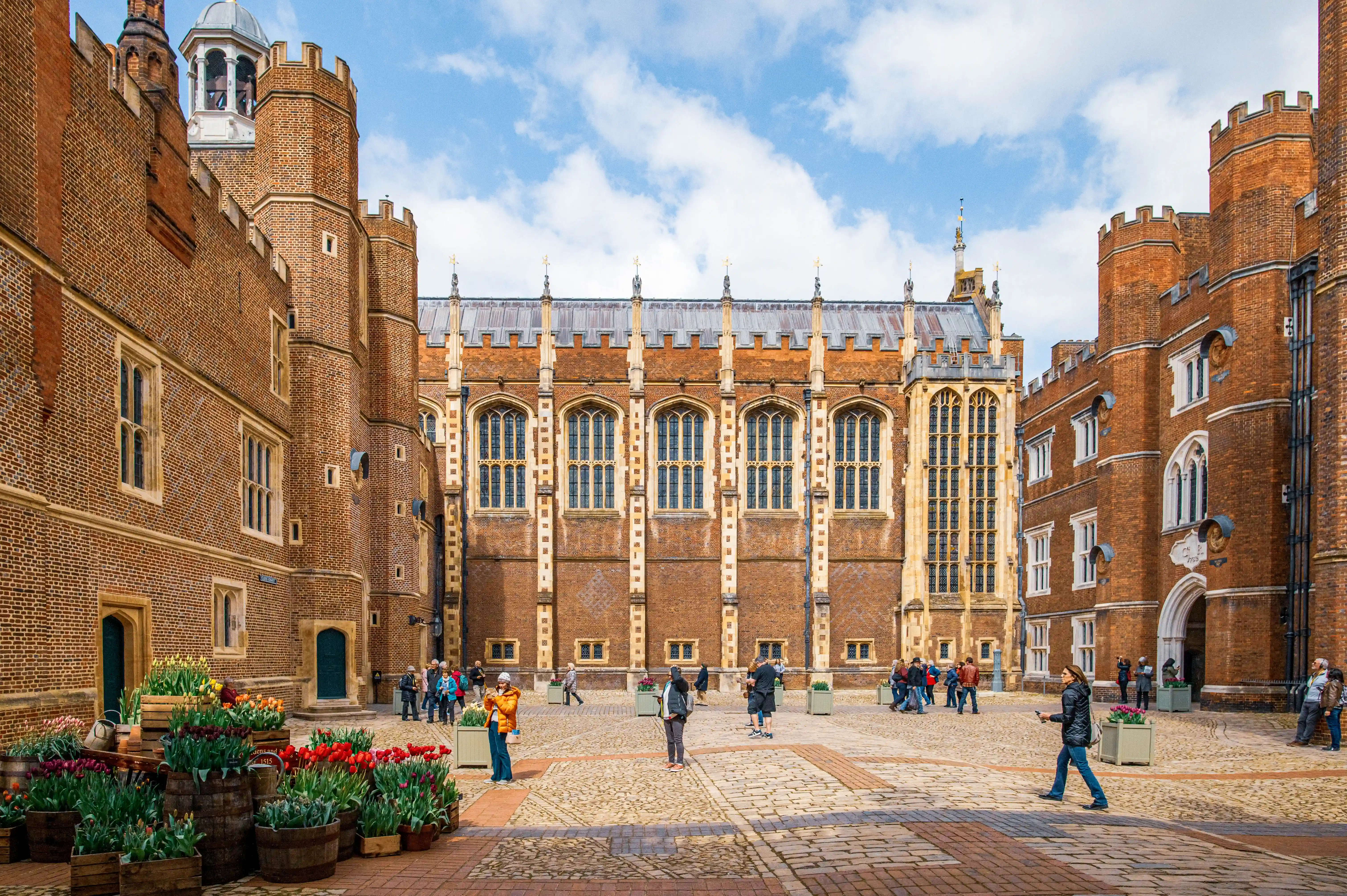 A wide-angle view of a courtyard at Hampton Court Palace. The courtyard is paved with brick and features large displays of red and pink tulips in wooden barrels and planters. Two impressive brick buildings with tall, arched windows and Tudor-style turrets flank the courtyard. In the background, a grand hall with a long row of windows and a castellated roof is visible. Several people are scattered throughout the courtyard, some admiring the flowers and others walking. The sky above is blue with scattered white clouds.