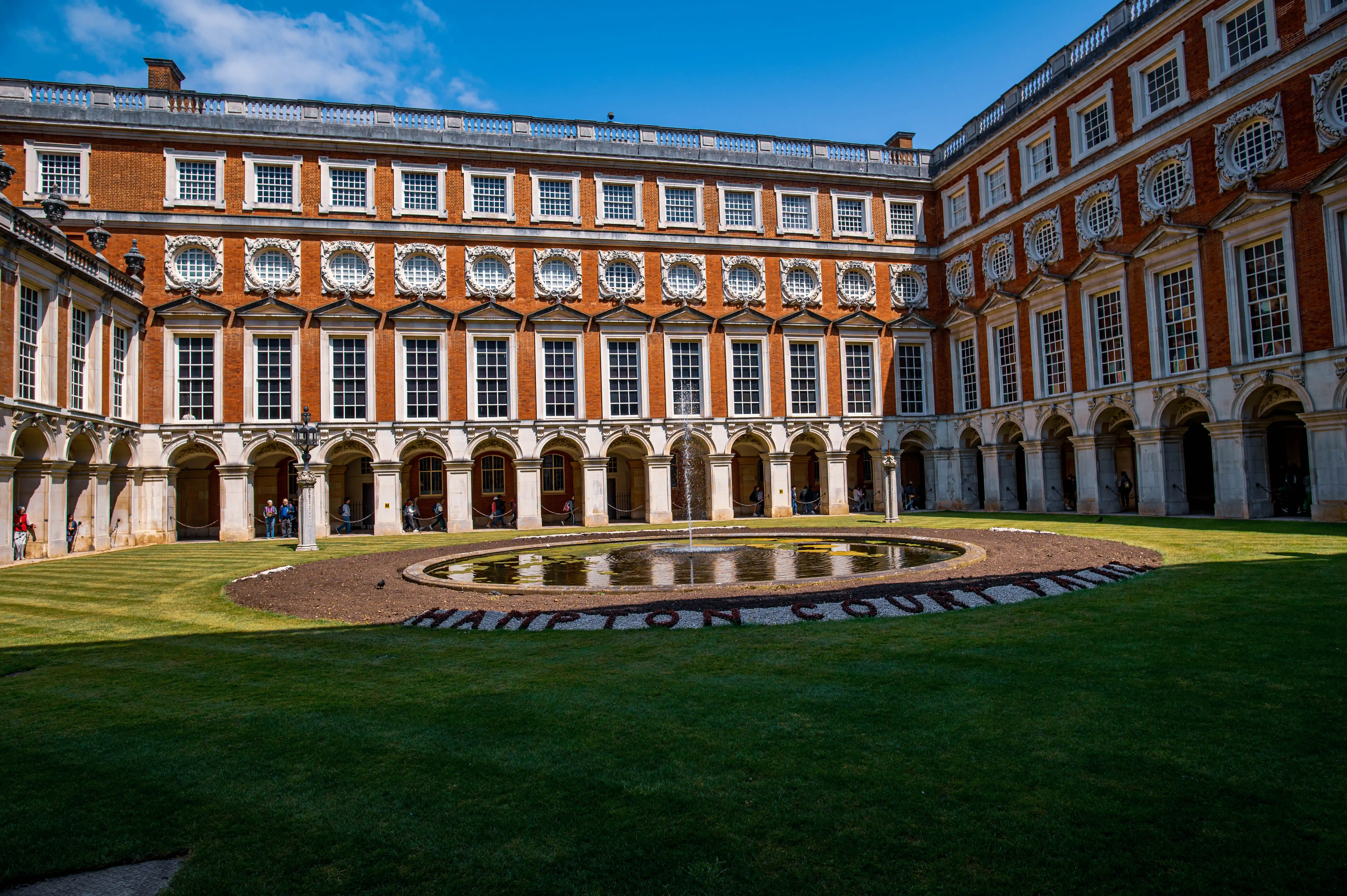 A sunlit courtyard of a historic palace with red brick buildings, white-trimmed windows, arched colonnades, a central circular fountain, and manicured green lawns.