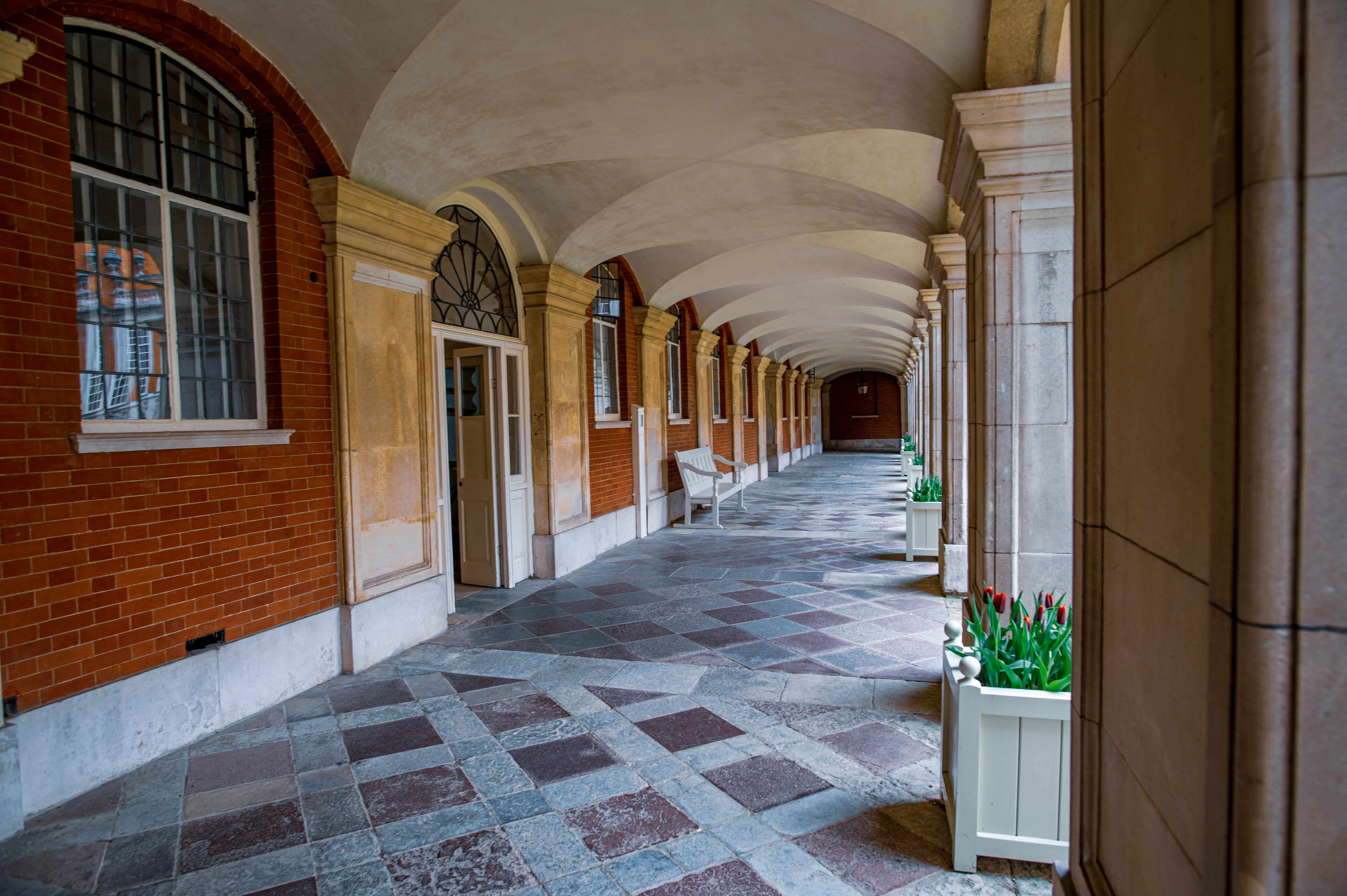 A symmetrical view down a stone-floored cloister walkway, with arched stone columns on the right and red brick walls with windows on the left. White planters filled with red tulips add a touch of colour.