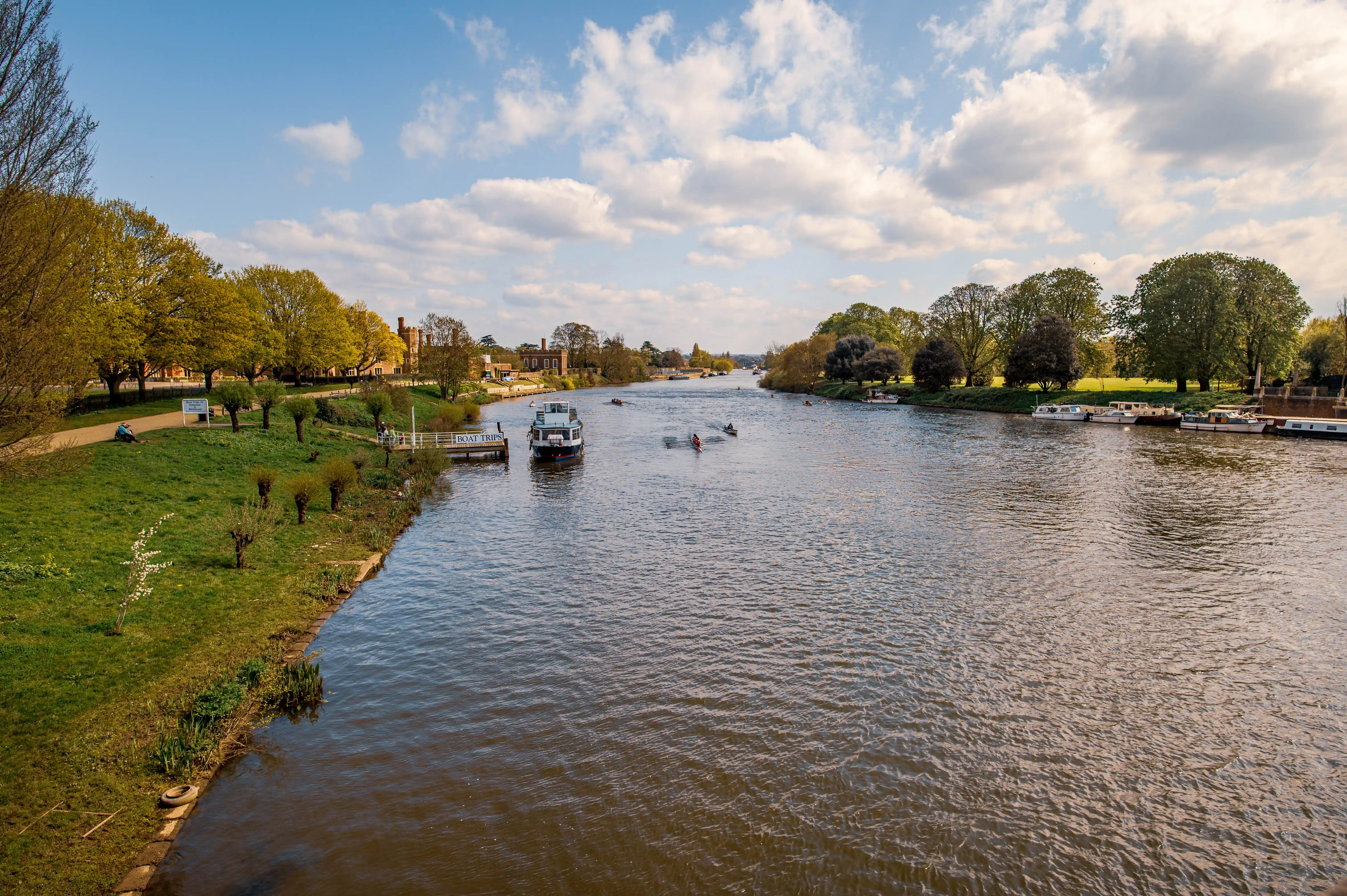 A wide river flows through a green landscape with trees and buildings on the banks. A boat is docked on the left, and kayakers are paddling on the water. Houseboats are moored on the right bank.