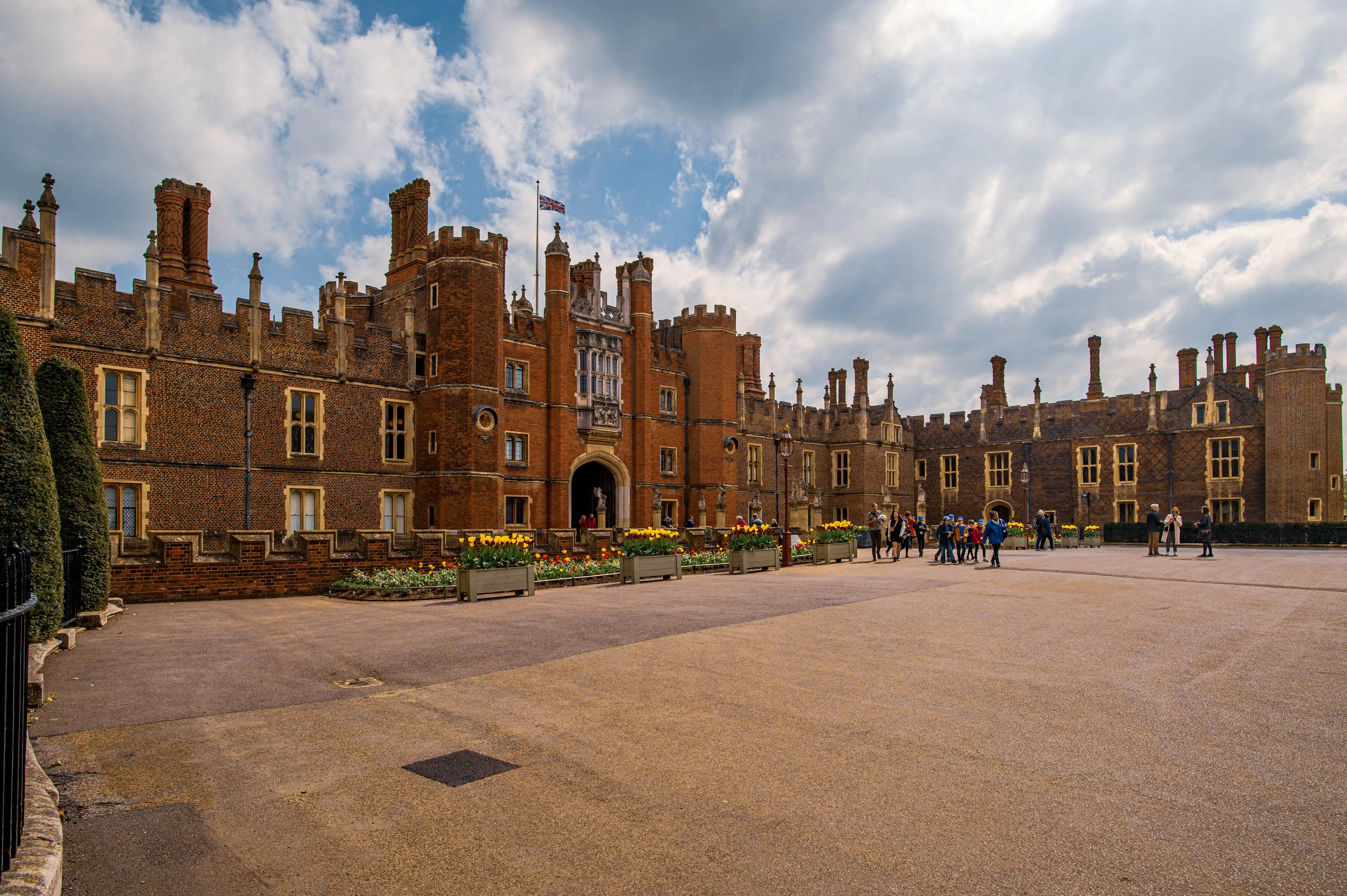 View of Hampton Court Palace with its grand brick facade, crenellated battlements, and numerous chimneys. Yellow flowers are planted in long rectangular boxes in front of the building, and a group of people are walking in the central courtyard.