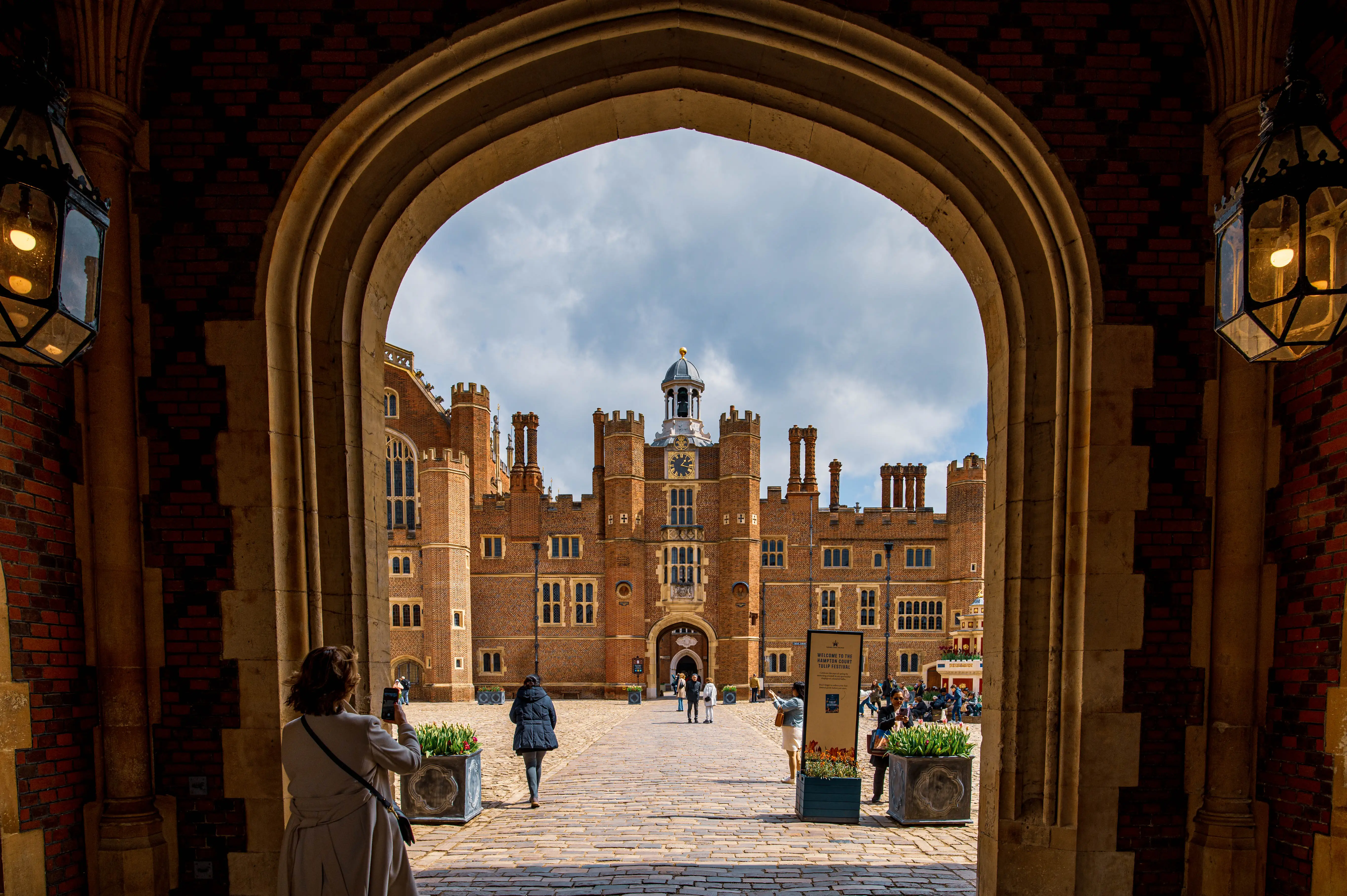 View of Hampton Court Palace's clock tower and facade through a large stone archway, with a woman on the left taking a photo and other visitors in the courtyard.