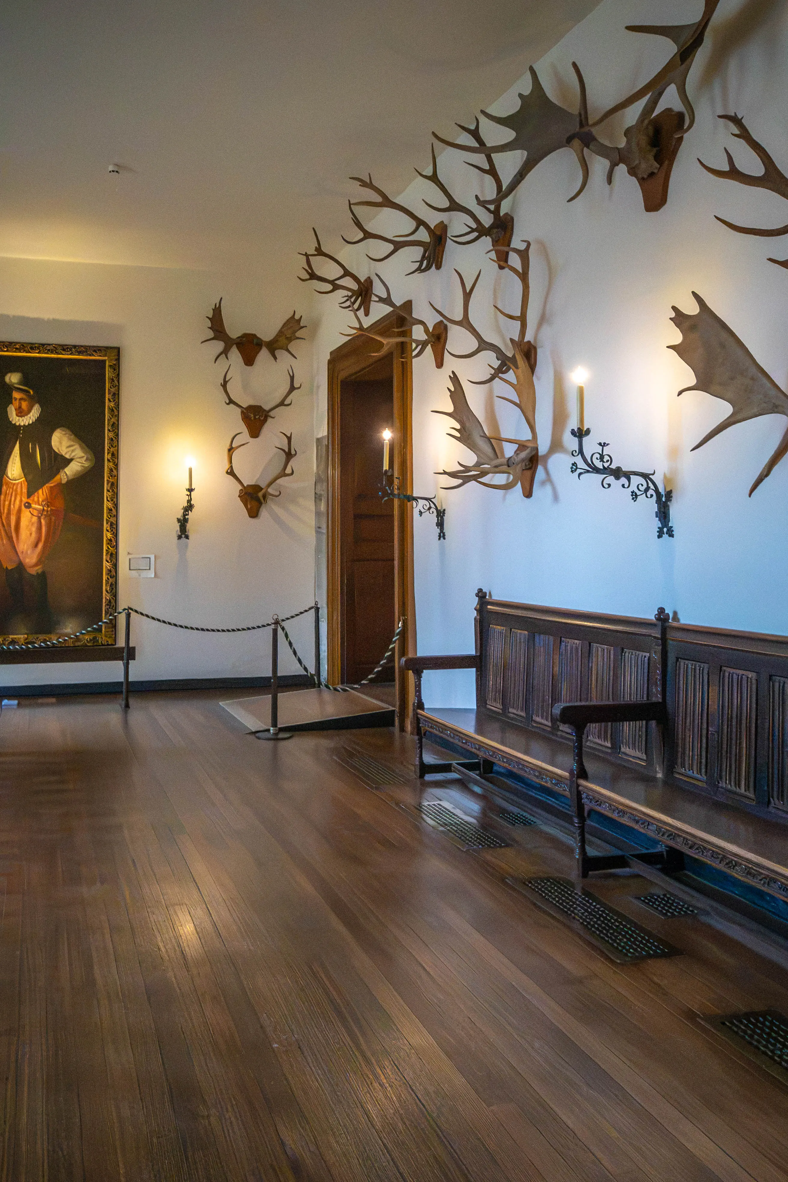 Historic gallery with mounted antlers, candle sconces, framed portrait and carved wooden bench.