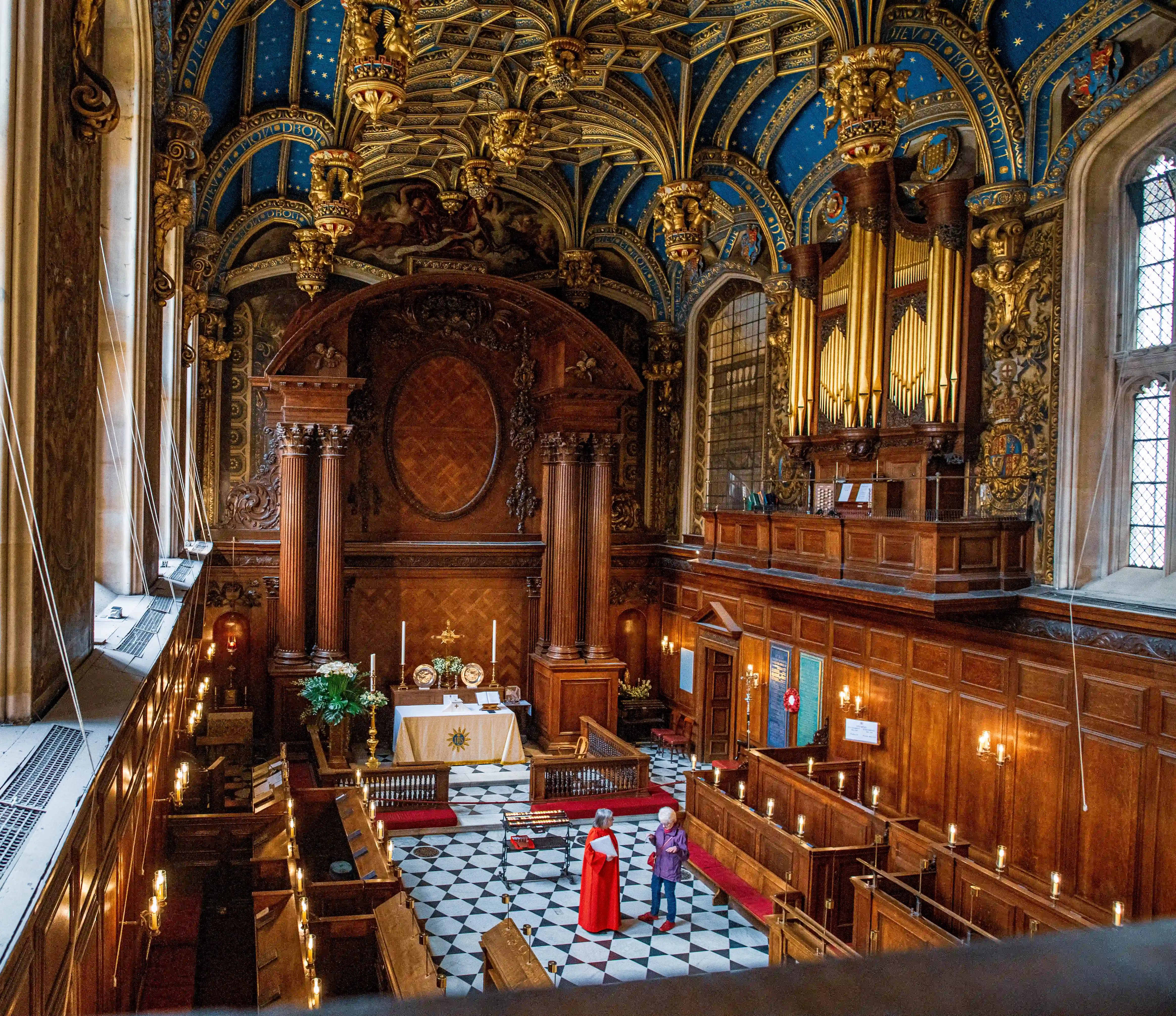 Ornate chapel with blue-and-gold vaulted ceiling, carved woodwork, altar, organ loft and two robed figures.