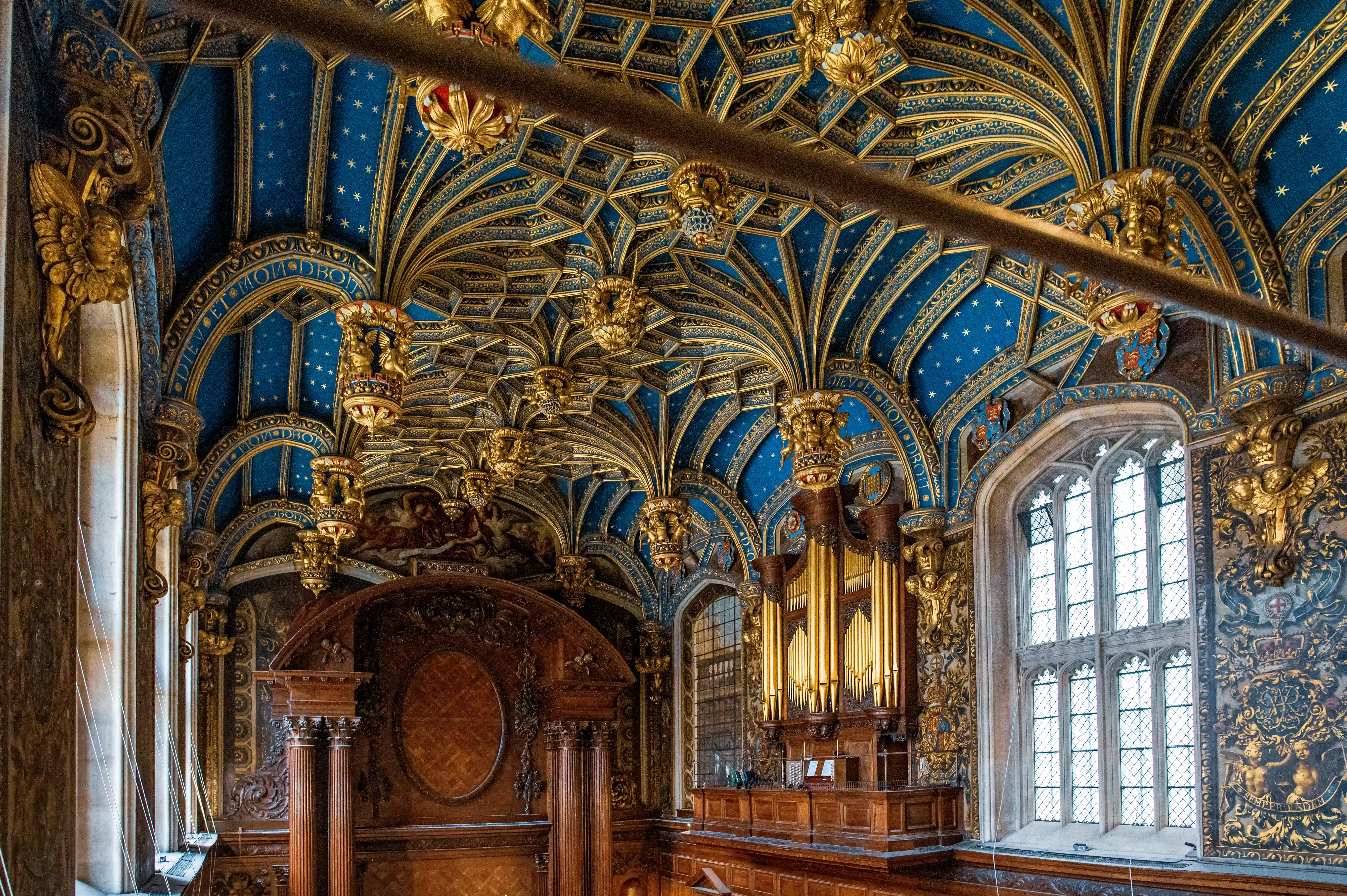Ornate chapel with blue-and-gold vaulted ceiling, gilded bosses, carved wooden structure, organ with golden pipes and tall multi-paned window.